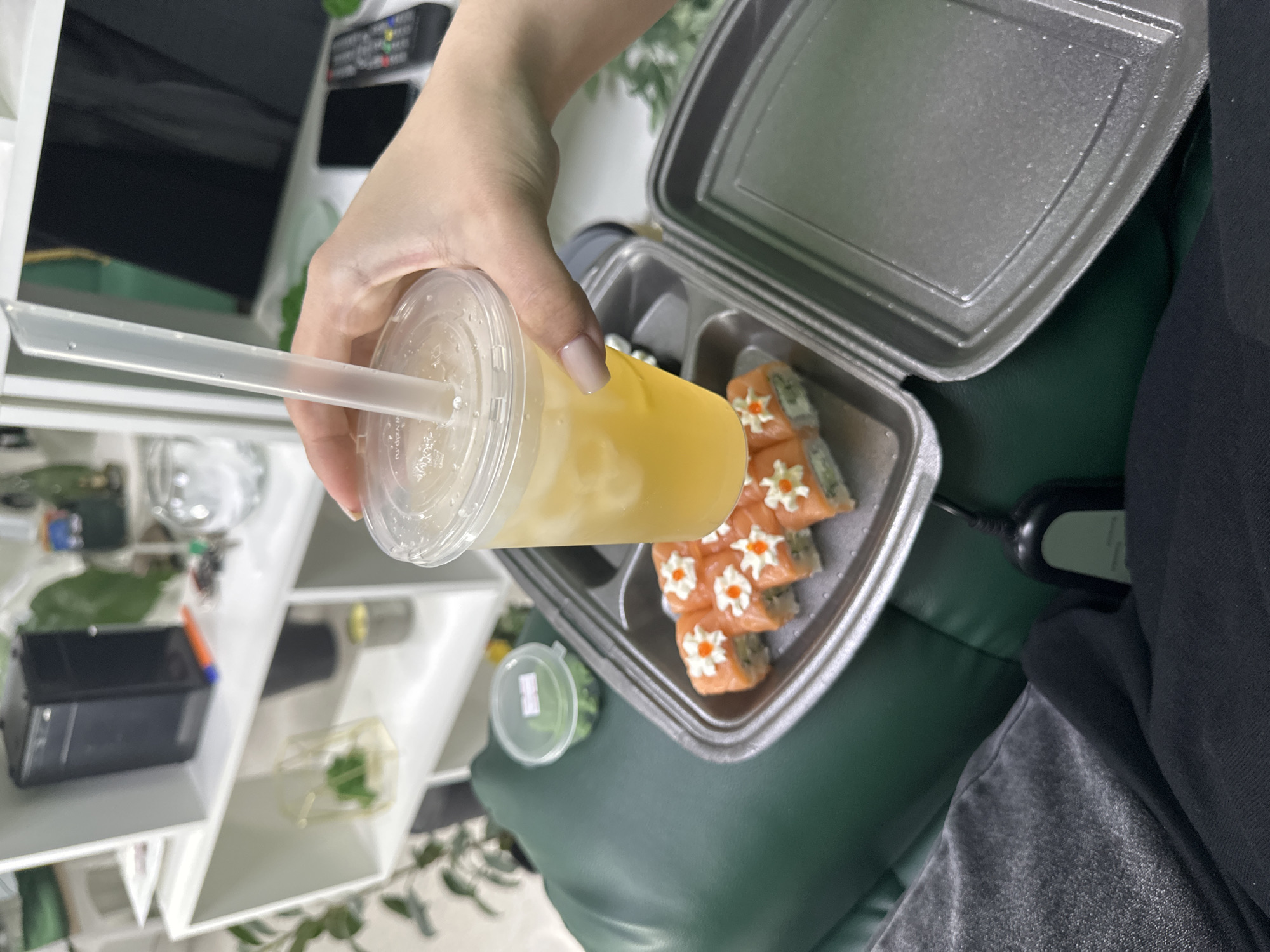 A human hand holds a plastic cup filled with a yellow liquid and a straw over a black plastic container of sushi on a green surface. The sushi rolls are orange with white decorations. A small container of green herbs is also present. The scene is inside, with a white shelving unit visible in the background, containing various items. No specific location or city can be identified from the image.