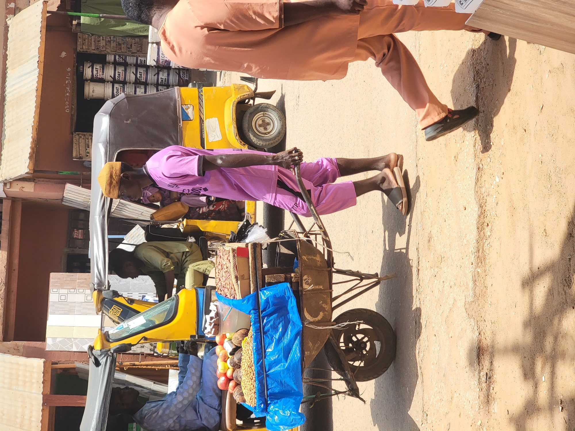 In Maiburiji, Nigeria, a man in a pink outfit and yellow cap pushes a cart piled with goods down a dirt road. The cart is covered with blue plastic and contains items such as peanuts and other foods. The road is near a building with paint cans visible. A yellow taxi-like vehicle is parked on the side of the road. A man in an orange outfit walks by.