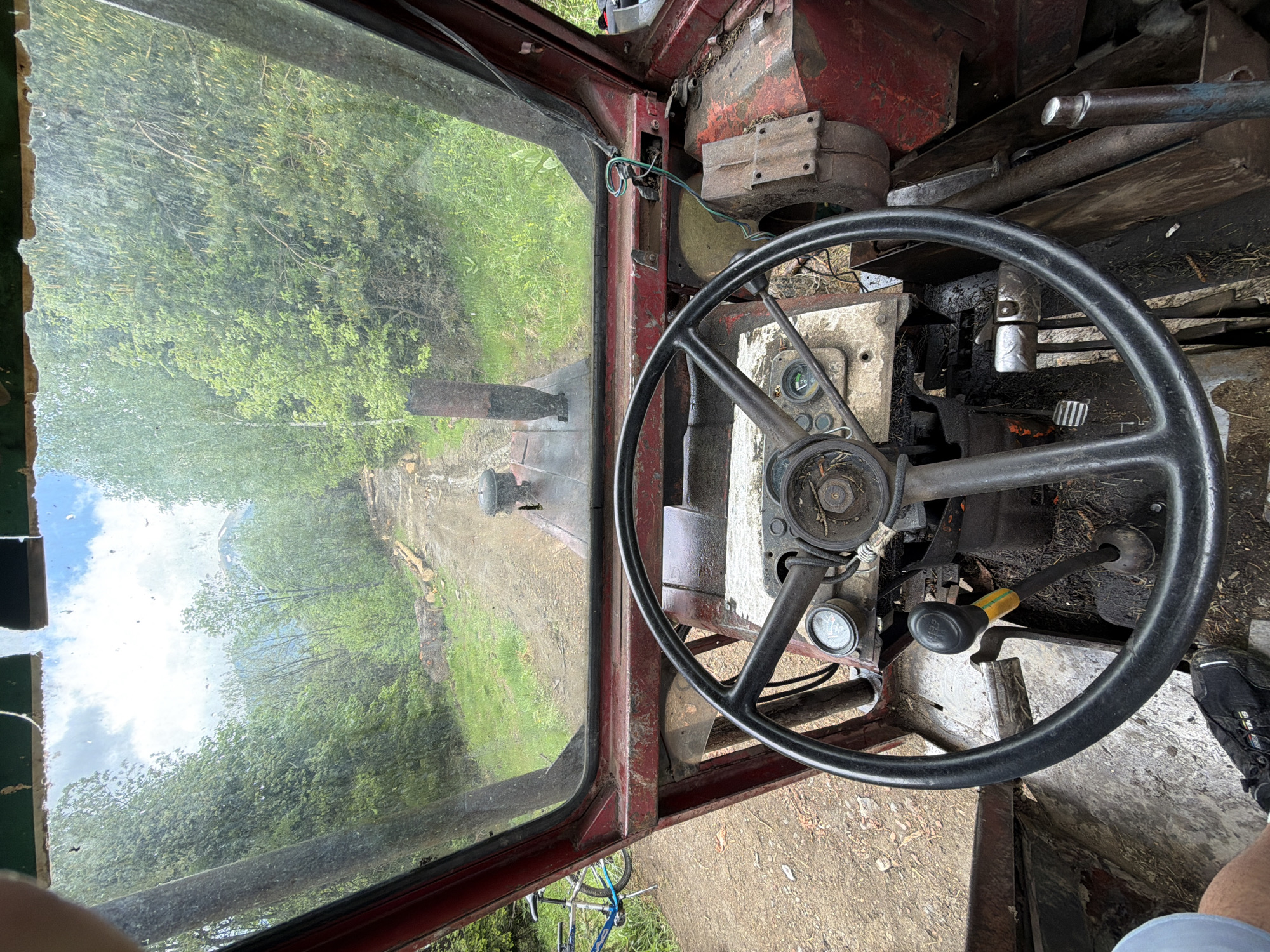 The image is an interior view from the driver's seat of a vehicle, possibly a tractor or a similar type of machinery. The environment outside the vehicle shows a rural area with a dirt road or path surrounded by trees and vegetation. The cabin of the vehicle is rudimentary, with a large steering wheel dominating the foreground. Various gauges and levers are visible on the dashboard. The vehicle appears to be located in a wooded area in the city of Рилски манастир, Bulgaria.