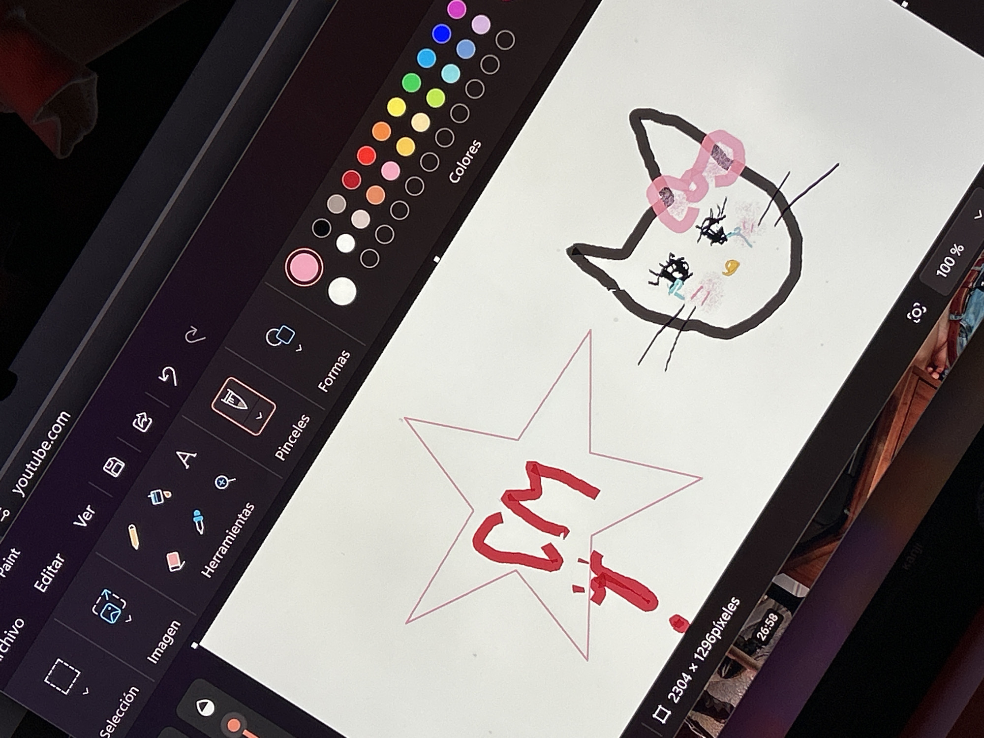 The image shows a close-up of a digital tablet screen displaying a drawing application. On the screen, a drawing of Hello Kitty is visible with a pink bow, black outline, and some details in different colors. Below the Hello Kitty drawing, there is a pink star with the letters "CM" written in red inside it. The application's interface is visible on the left side of the screen, showing various tools and color palettes. The bottom left shows the word "youtube.com" and on the bottom right it shows "2304 x 1296 pixeles" and "26:58". A glimpse of a wooden surface and a person's leg can be seen at the edge of the tablet screen. There is no indication or information given that this image is located in Buenos Aires, Argentina.