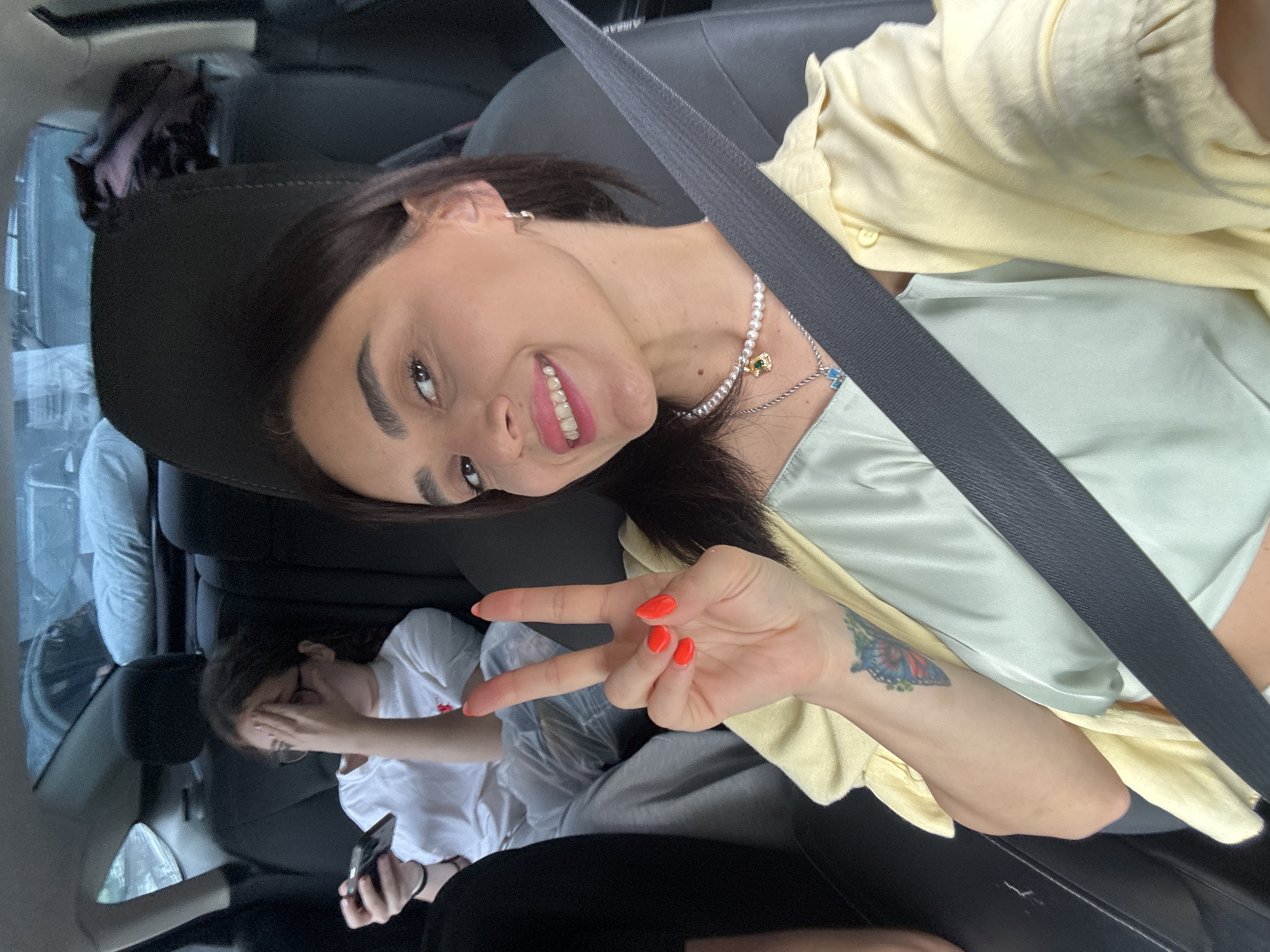 The image shows a selfie taken from inside a car, likely in Tbilisi, Georgia. The main subject is a woman in the front passenger seat, smiling and making a peace sign with her right hand. She is wearing a light yellow button-up shirt over a light green slip dress. She has on a pearl necklace with a pendant and hoop earrings. A tattoo of a colorful butterfly is visible on her left forearm. In the back seat, another person, a woman wearing glasses and a white t-shirt, is visible looking at her phone. The interior of the car is visible, including the headrests and seatbelts. The background appears to be a street scene, possibly with other vehicles. The lighting suggests it is daytime.