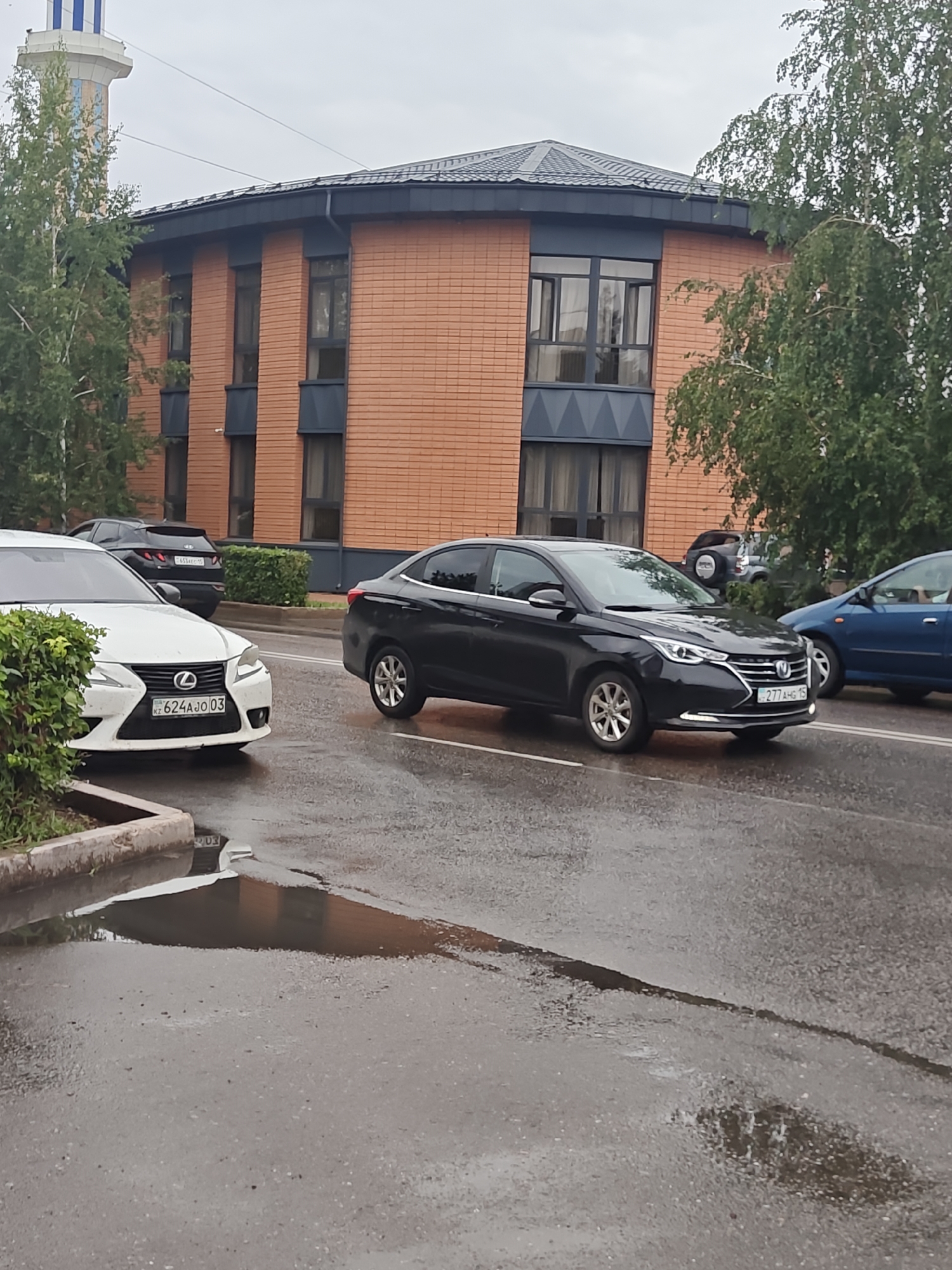 The image depicts a street scene in Petropavl, Kazakhstan, on a cloudy day, with wet asphalt indicating recent rain. In the foreground, a white Lexus is parked partially to the left, its license plate visible as KZ 624AJ0 03. To the right of the Lexus, a black sedan is driving on the wet road. Further back, another black SUV is visible, its license plate partially obscured. A blue car is also on the road to the right of the black sedan. In the background, a modern building with orange brick facade and dark grey trim and roof stands prominently. To the far left, a tall, slender structure, possibly a water tower, with blue and white banding is partially visible. Trees with green foliage are present on both sides of the street. The overall environment suggests a typical urban setting.