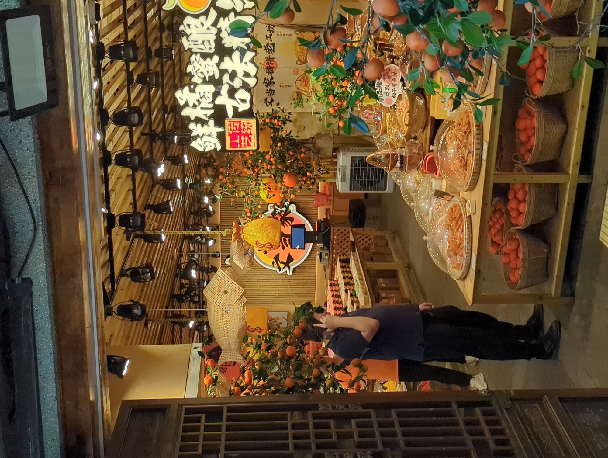 The image depicts a street-side fruit stall, likely located in the Yuzhong District of China, characterized by its traditional Chinese architectural elements. The stall is laden with fresh produce, primarily oranges and other citrus fruits, displayed in woven baskets and on tiered wooden shelves. Decorative elements, such as fish-shaped lanterns and hanging bags, suggest a festive atmosphere or a special promotion.
A man, dressed in a dark blue polo shirt and black trousers, is prominently featured in the foreground, talking on his mobile phone. He is standing near a display of oranges, his posture suggesting a casual conversation. Behind him, another individual, wearing an orange t-shirt and black trousers, is visible, partially obscured by a fruit tree.
The stall's backdrop features Chinese characters, likely advertising the fruits or the establishment. The lighting is a mix of overhead lamps and ambient light, creating a warm and inviting scene. A white portable fan is placed near a display of various food items, possibly dried fruits or snacks, indicating that the stall also offers processed goods. The overall impression is one of a bustling marketplace, offering fresh produce and local delicacies in a vibrant setting.