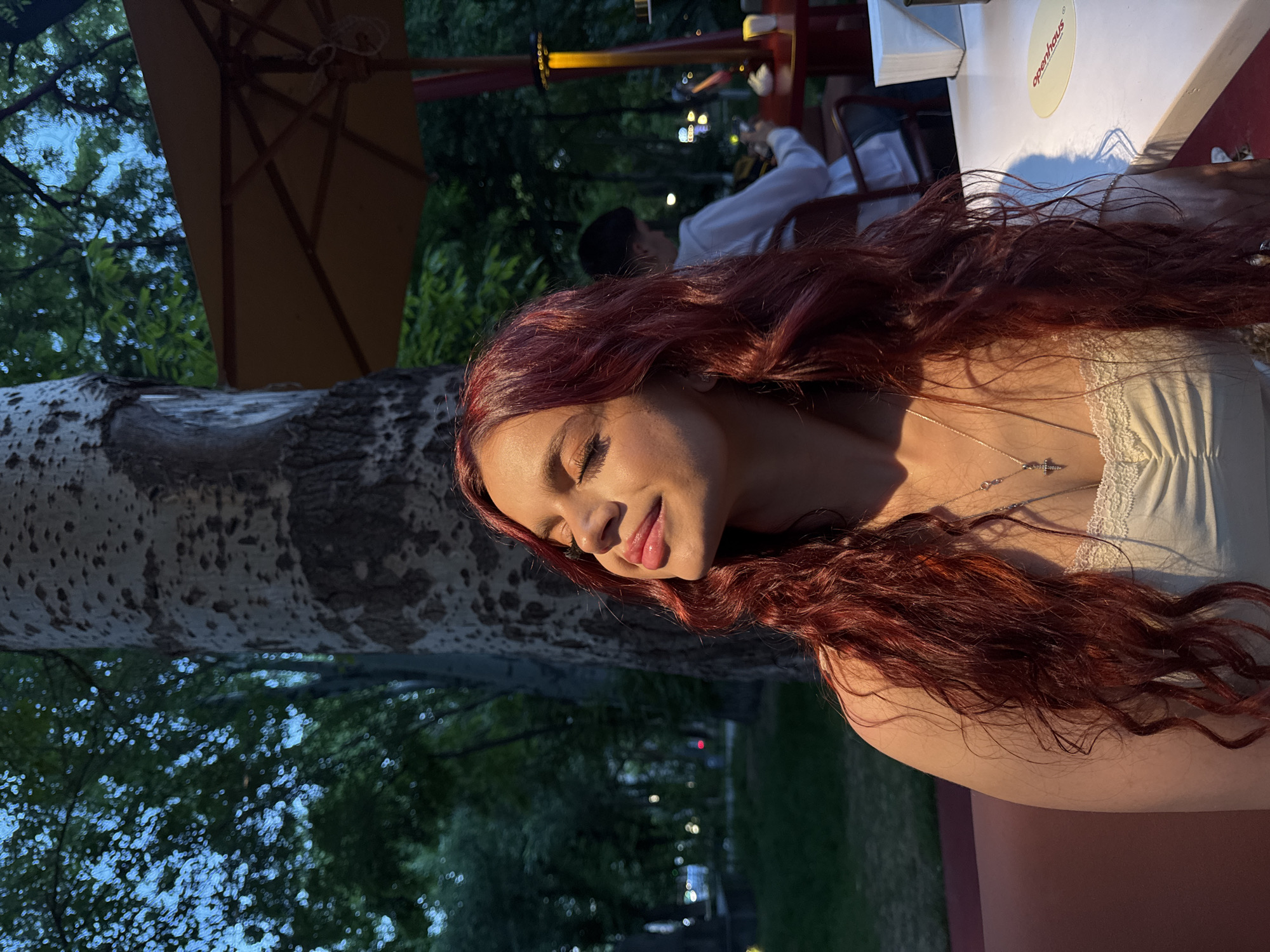The image portrays a young woman with long, wavy, reddish-brown hair, captured in an outdoor setting during the golden hour or early evening. She is seen in profile, with her eyes gently closed and a soft smile, conveying a sense of peace or contentment. Warm, directional sunlight illuminates her face, shoulder, and flowing hair. She is wearing a light-colored top with a delicate lace trim and a silver necklace featuring a small cross pendant.
To the left, a large tree trunk with mottled bark is prominent, and above it, a brown patio umbrella is partially visible, indicating an outdoor dining area. In the blurred background, another person, possibly a man in a white shirt, is seated at a distant table. The right side of the image features a white table surface where a circular label with the text "openhaus" is visible in red, possibly with "studio" underneath. A faint shadow, likely cast by the woman's hair and arm, stretches across the table. Numerous bokeh lights in the deep background suggest artificial illumination or distant city lights, confirming the evening ambiance. The overall scene depicts a relaxed outdoor moment, potentially at a cafe or restaurant in Yerevan, Armenia.