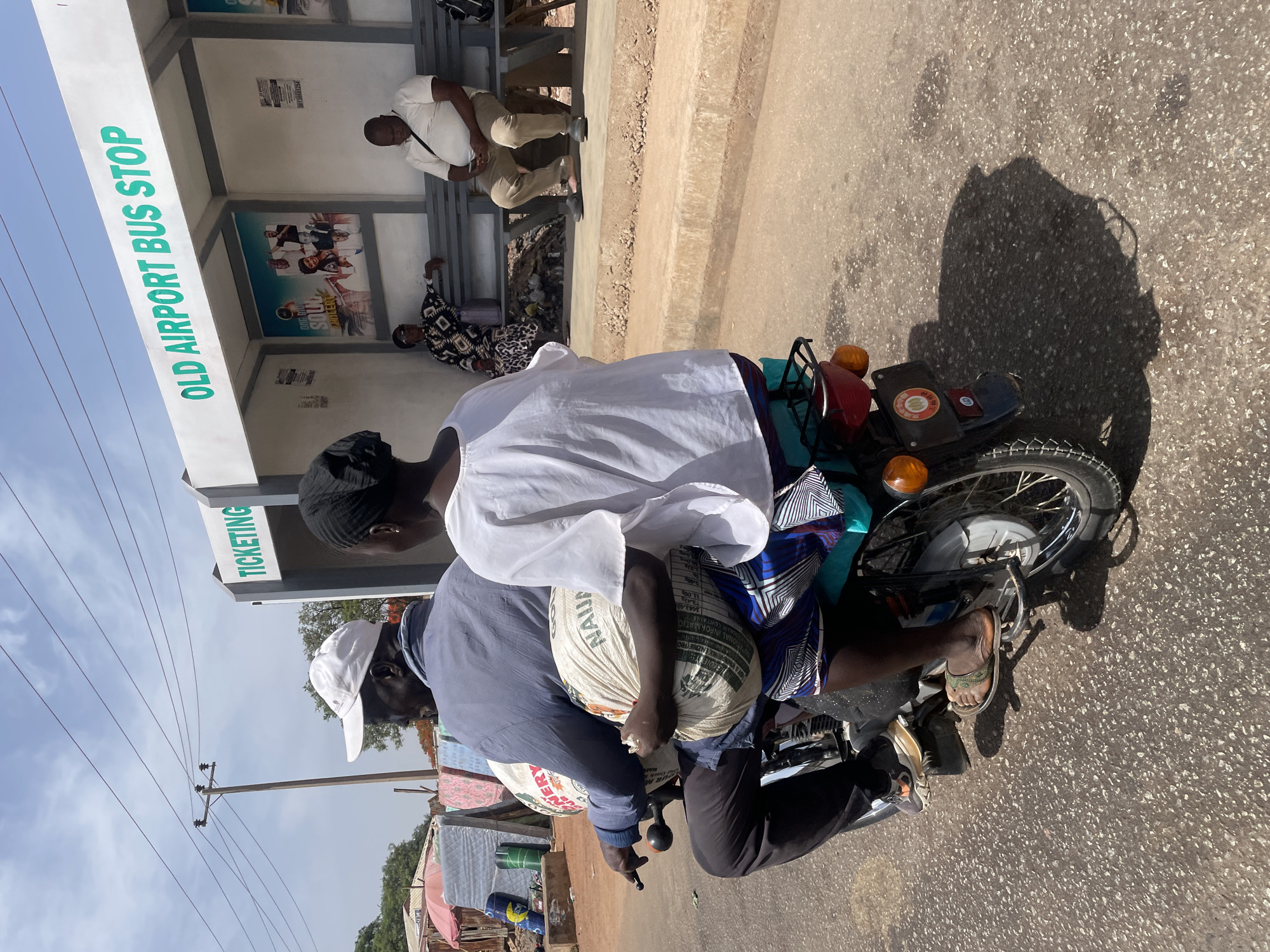 A medium, eye-level shot shows two people on a motorcycle stopped near the Old Airport Bus Stop in Jos, Nigeria. A man in a grey shirt and white cap is driving the motorcycle. He is holding a large sack of grain, labeled "NAJA", in front of him. Behind him, a person in a white shirt and black head covering is seated. The motorcycle is parked on the side of a paved road. The Old Airport Bus Stop is visible in the background, with people sitting on benches inside. A sign above the bus stop reads "OLD AIRPORT BUS STOP" and "TICKETING". Power lines and trees are visible in the distance, alongside buildings on the roadside.
