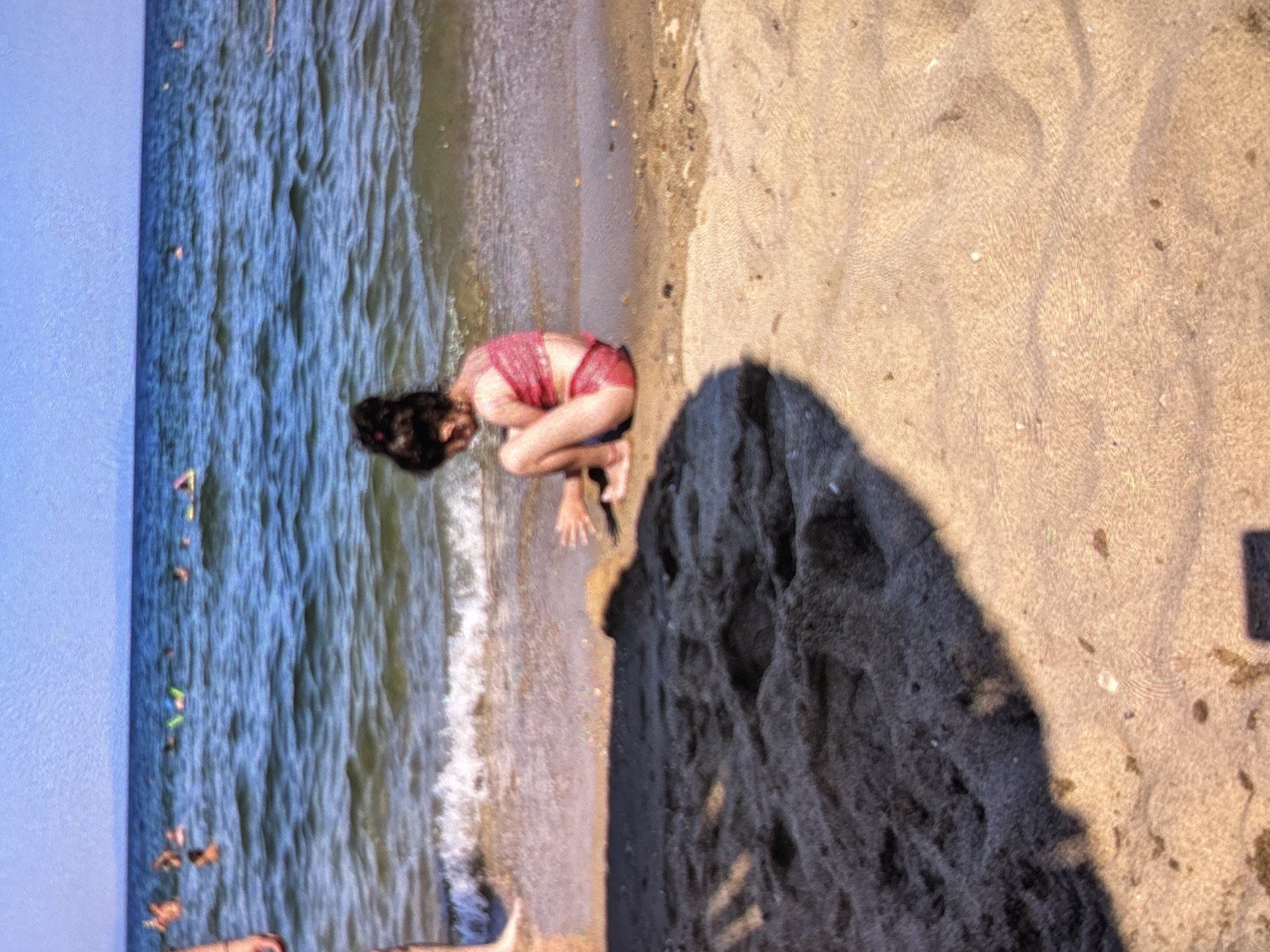 A bright, daytime image captures a beach scene. A person, appearing to be a child or young adult, is seated or kneeling on the wet sand at the edge of the water. They are wearing a red or pink swimsuit and have dark hair that partially covers their face as they look down. The water, a calm blue-green, gently laps at the shore. Further out in the water, several small, colorful objects and indistinct figures, likely other people, are visible, suggesting other activity. The sand is a light tan color, and a large, dark shadow falls across the dry sand in the upper right portion of the frame, indicating bright sunlight, possibly during the late morning or afternoon. The overall setting is a sunny beach. There is no visible text.