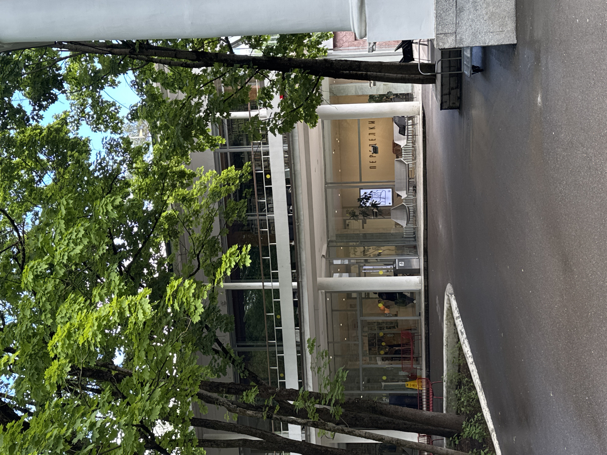 A ground-level photograph shows an exterior view of a modern building behind a lush tree. The building is primarily glass and white metal, with the name "PEREDELKI" visible above the entrance. Light is coming in through the glass walls and highlighting the interior.
The surrounding environment includes a paved area that appears wet, and a dense green tree with leaves framing the building. The photograph seems to be taken in Vnukovo, Russia, based on the name of the building. There are no apparent people visible.