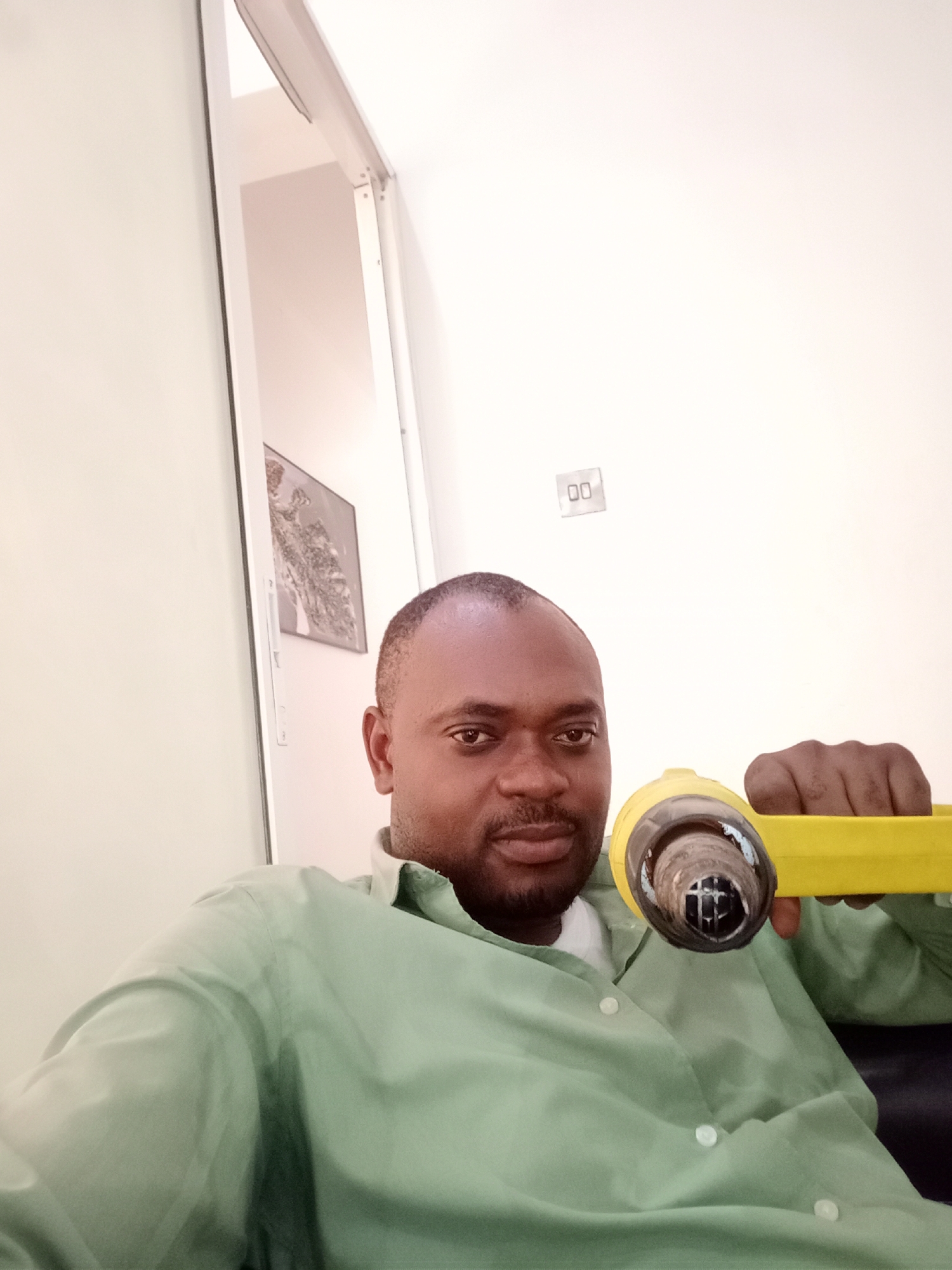 A man, likely in his 30s or 40s, with dark skin, short dark hair, and a beard, is featured in a close-up indoor photograph taken in Gwarinpa, Nigeria. He is wearing a light green collared shirt over a white undershirt and looks directly at the camera with a neutral expression.
He holds a bright yellow tool, possibly a heat gun or a similar device, with its dark, circular nozzle pointed towards the camera. The background consists of plain, light-colored walls, suggesting a simple indoor setting such as a home or office. To the left, a partial view of an open doorway or cabinet reveals a glimpse of a framed picture on an inner wall. On the right wall, above the man's head, a white electrical light switch or outlet with two square slots is visible. The bright lighting indicates it is daytime. There is no visible text in the image.