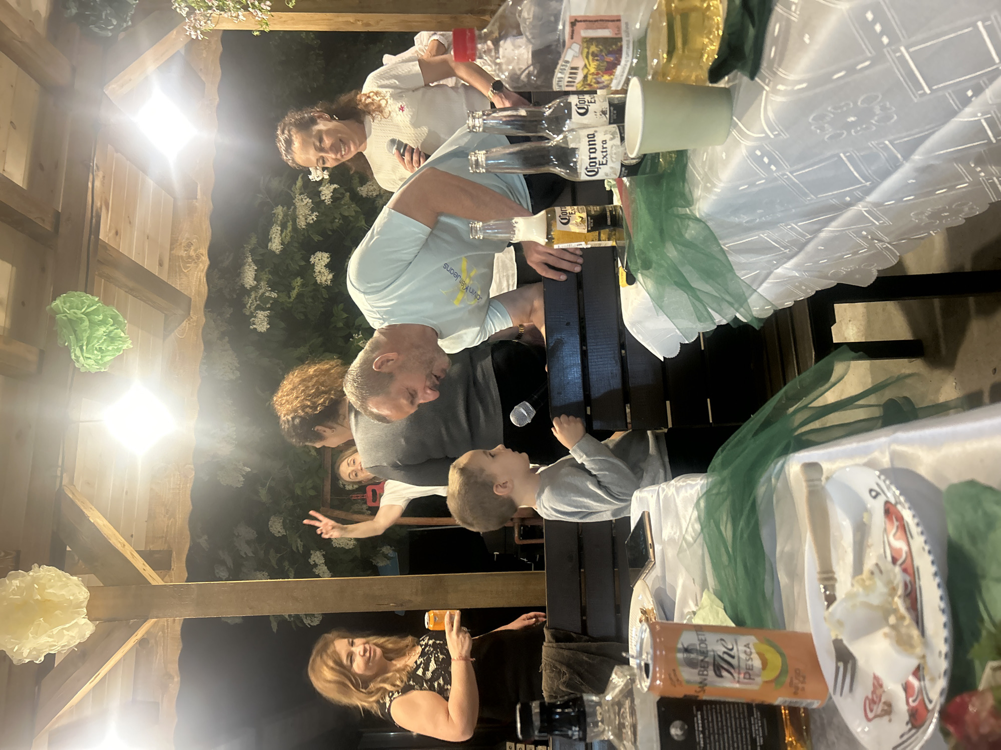 The image shows an outdoor gathering. Several people are present, including adults and a child. A table covered with a patterned white tablecloth holds items such as Corona Extra beer bottles, a paper cup, and a can of San Benedetto iced tea. There is a plate with food on it. People appear to be talking and interacting. The gathering takes place under a wooden structure decorated with paper pom-poms and greenery, possibly at night, illuminated by lights. While the information mentions Sandrovo, Bulgaria, the image itself does not provide enough data to pinpoint the precise location within Sandrovo.