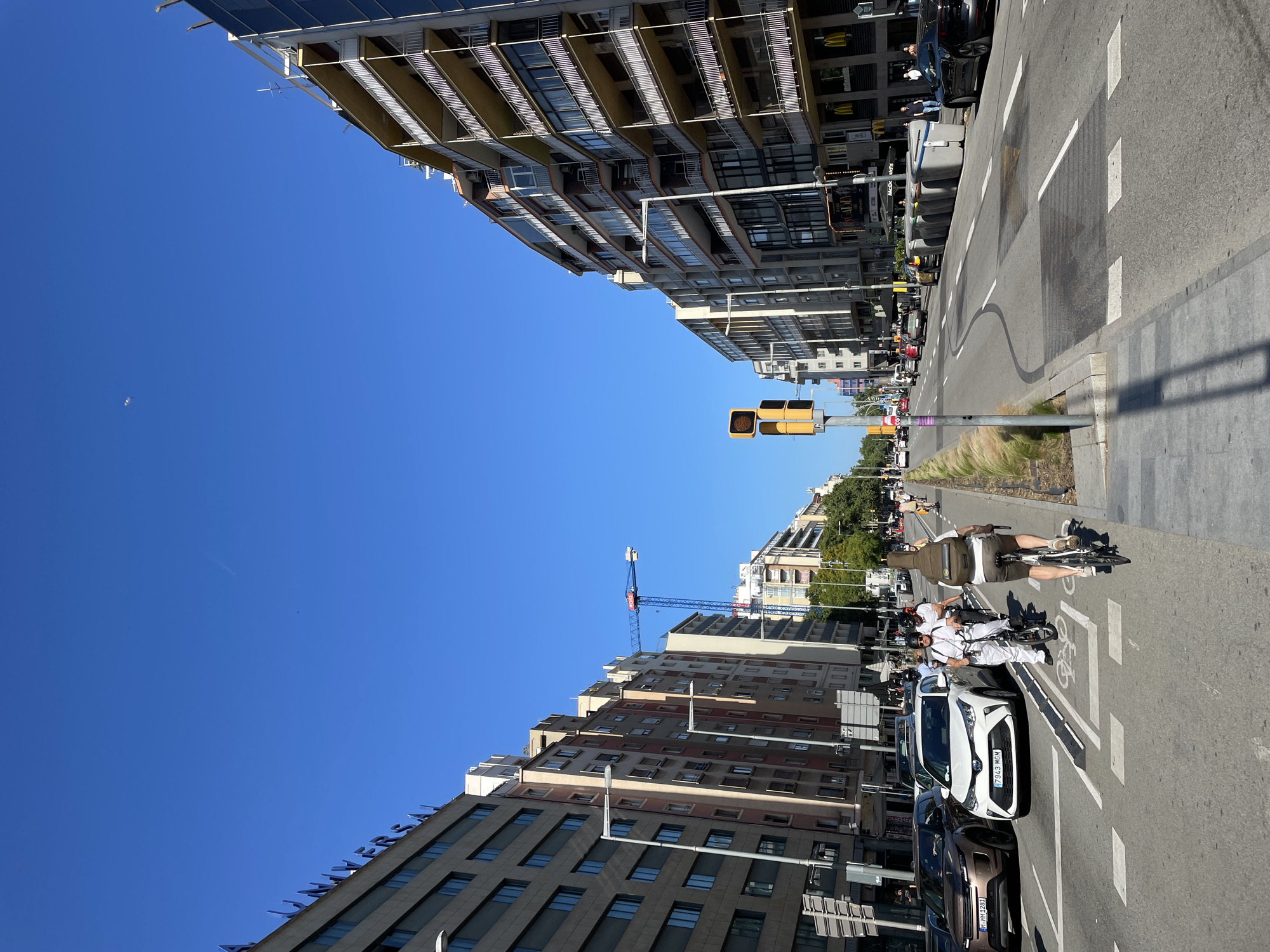 This image captures a vibrant, sunny street scene in an urban area of Barcelona. The photograph is taken from a slightly elevated perspective, looking down a wide thoroughfare flanked by multi-story buildings under a clear blue sky.
On both sides of the street, tall residential and commercial buildings dominate the view, featuring numerous windows and balconies. The building on the left (when correctly oriented) has part of the word "UNYVERSAL" visible on its facade, while buildings on the right show signs for "McDonald's" and "GODIVA". Some buildings also display solar panels on their rooftops. A construction crane is visible in the distance, indicating ongoing development.
The street itself is bustling with activity. It has several lanes, including a dedicated bicycle lane marked with white symbols on the asphalt. Various vehicles are present: cars are parked along the curb and others appear to be in motion. A white car with license plate "7943 MGM" is prominently featured. There are also several people on bicycles and electric scooters, one cyclist carrying a large instrument case on their back. Yellow traffic lights and streetlights line the road, alongside patches of green bushes acting as a median or divider.
The scene depicts an ordinary daytime city environment, characterized by bright sunshine and a cloudless sky, suggesting pleasant weather conditions. No specific emotions are discernible, portraying a typical day in the city. Other visible text includes "M 1261" on another license plate.