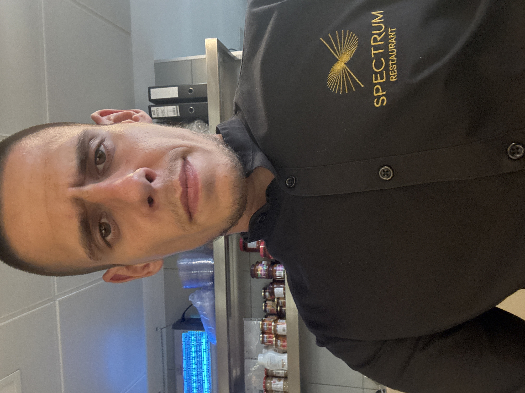 A man in a black chef's uniform with the "Spectrum Restaurant" logo embroidered in gold on the right side of his chest is posing for a selfie. He has short hair, a light complexion, and is looking directly at the camera with a neutral expression. The background appears to be a commercial kitchen, with shelves stocked with jars and containers, and what looks like a commercial fly zapper on the top shelf. The overall impression is a candid, informal photograph taken in a workplace. The location is not specified in the image.
