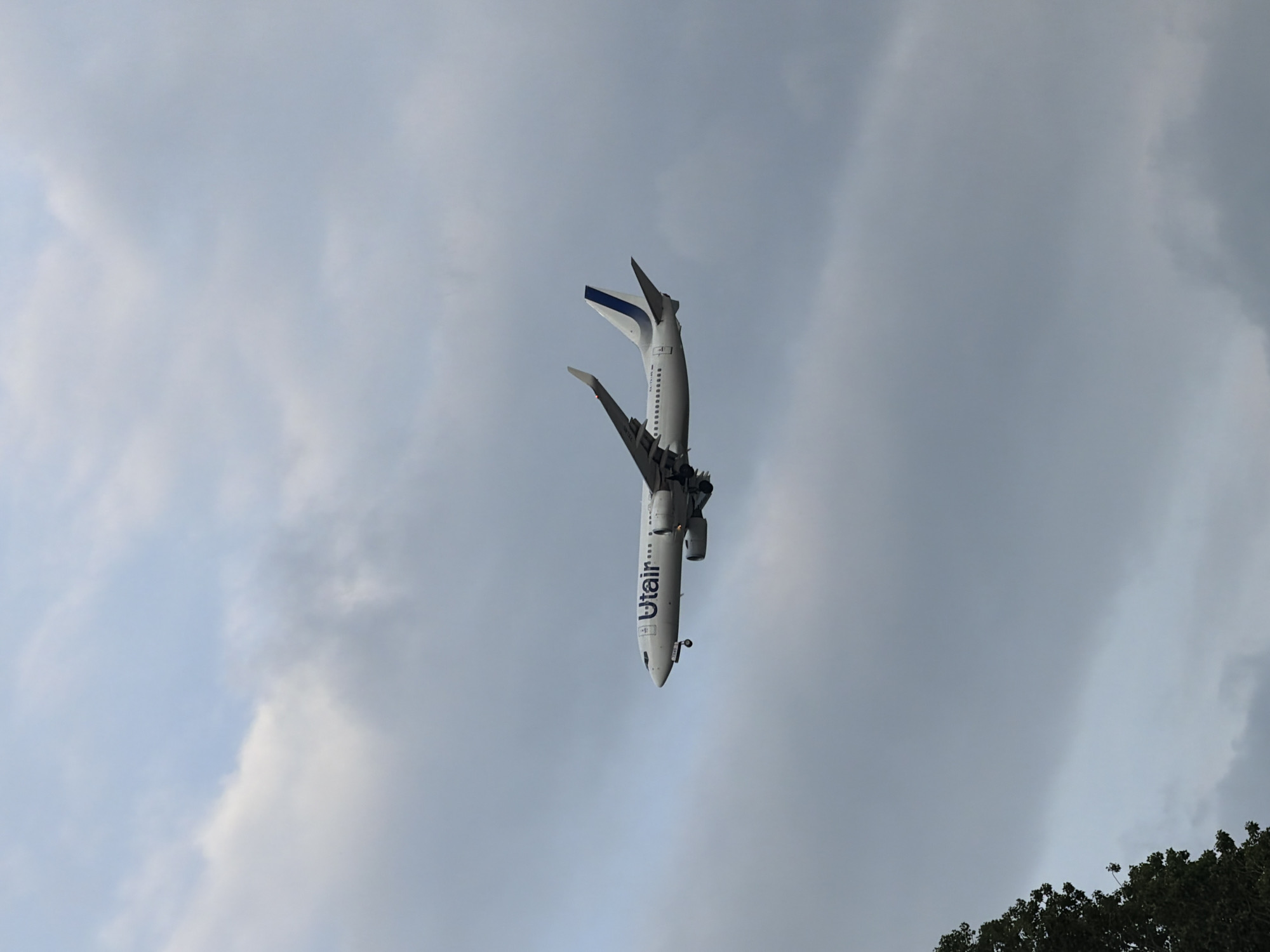 This is a low-angle shot of an airplane flying in the sky. The airplane is upside down, with its nose pointing downwards. The fuselage is white with blue lettering that reads "Utair". The wings are also white with blue accents on the wingtips. The underside of the airplane is visible, showing landing gear and engines.
The sky is overcast with a mix of light and dark gray clouds. The lighting suggests it is daytime, possibly late afternoon or early evening, given the diffused light and the presence of some darker clouds. The bottom right of the image shows a dark green tree line, suggesting the photograph was taken from below and somewhat close to the ground. The location context provided is Sochi, Russia, which implies a possible coastal or mountainous environment given the area. No people are visible in the image. The overall mood is somewhat dramatic due to the unusual orientation of the airplane against the cloudy sky.