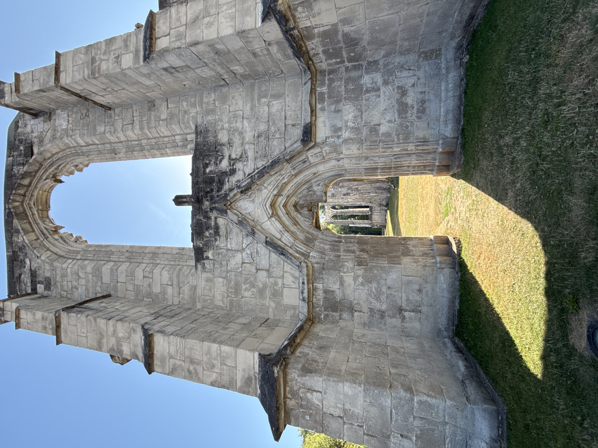 The image shows the ruins of the Walkenried Abbey in Walkenried, Germany. The ruins are made of light-colored stone and are partially overgrown with grass and other vegetation. The sky is clear and blue. The image is taken from a low angle, looking up at the ruins. In the foreground, there is a large arched doorway. Through the doorway, other ruined arches can be seen. The sun is casting long shadows across the grass.