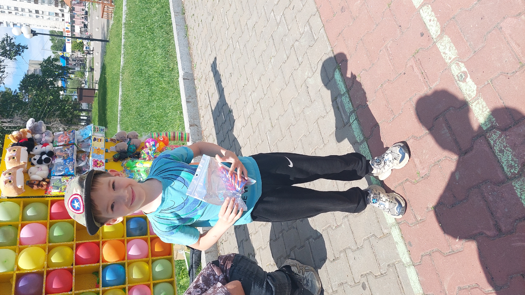 A young boy in a blue t-shirt and black athletic pants, wearing a camouflage baseball cap with a Captain America emblem, stands on a paved walkway, holding a clear plastic bag filled with small colorful toys or candies. He is smiling directly at the camera. Behind him, to his left, is a colorful carnival-style game booth with rows of balloons and hanging plush toys. To his right, there is a grassy area with trees, buildings, and streetlights, indicative of an urban park or public space. The scene suggests a sunny day at an outdoor event or fair. The architecture and streetscape in the background are consistent with Komsomolsk-on-Amur, Russia.