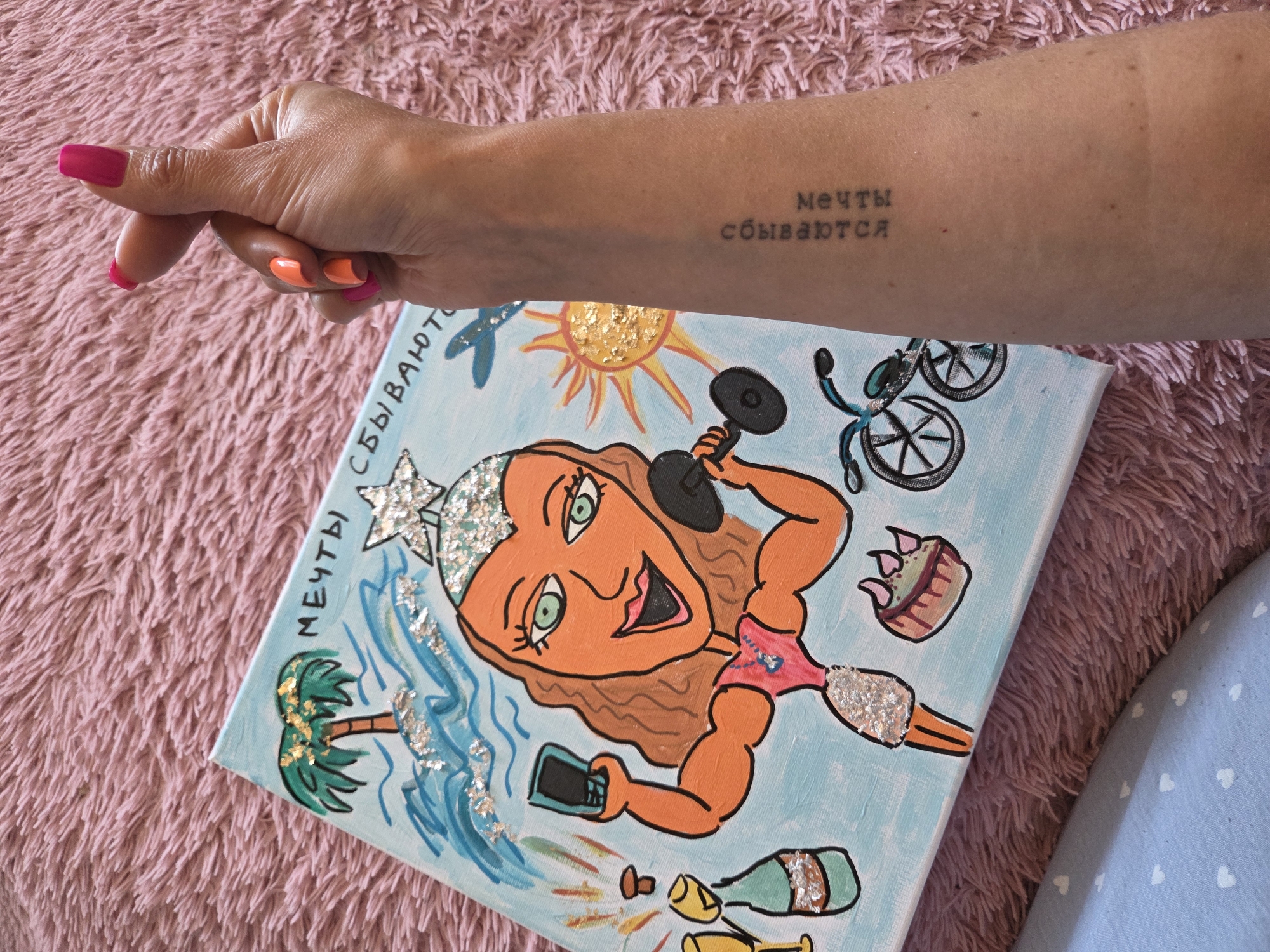 A woman's arm with a tattoo in Russian, "Мечты сбываются" (Dreams come true), is extended over a canvas painting lying on a pink, furry blanket. The woman's hand has nails painted in orange and pink. The painting depicts a cartoonish female figure with orange skin, brown hair, and green eyes, wearing a star-shaped crown. The figure is holding a dumbbell in one hand and a smartphone in the other. The painting also includes a palm tree, waves, a sun, a bicycle, a cupcake, and a champagne bottle. The canvas has the same phrase "Мечты сбываются" painted on it. The background of the painting is light blue, and elements of glitter or silver foil are incorporated into the design. The location is likely a personal residence rather than a public place in Birobidzhan, Russia, although this cannot be definitively stated.