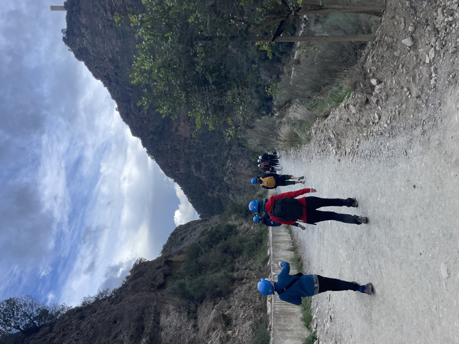 The image depicts a group of people hiking along a narrow dirt and gravel path winding through a mountainous gorge, characteristic of the landscape around El Chorro, Spain. Steep, rocky mountainsides covered with sparse vegetation and trees rise dramatically on both sides of the path, receding into the distance under a partially cloudy sky with patches of blue.
A line of at least eight individuals is visible, walking away from the camera. The subjects are dressed in outdoor attire, including jackets and pants, and most notably, they are wearing bright blue helmets and carrying backpacks, suggesting participation in an organized activity that requires head protection, such as trekking in a challenging or protected area. The person in the foreground wears a blue jacket, blue helmet, and a small black shoulder bag. Immediately ahead, another individual is seen in a red jacket, black backpack, and blue helmet. Further down the path, a person with a yellow backpack and blue helmet is visible among the group.
The activity taking place is a guided hike or trek through the mountainous terrain. The presence of safety helmets points towards an adventure tourism activity, possibly related to routes like the Caminito del Rey which is famous in the El Chorro region.
Notable details include the time of day, which appears to be daytime with natural light, likely late morning or early afternoon. The weather is partly cloudy but suitable for outdoor activities. In the mid-ground, a concrete structure, possibly part of an old irrigation channel or walkway, runs parallel to the path. No visible text is present in the image. The scene conveys a sense of adventure and exploration within a rugged natural environment.