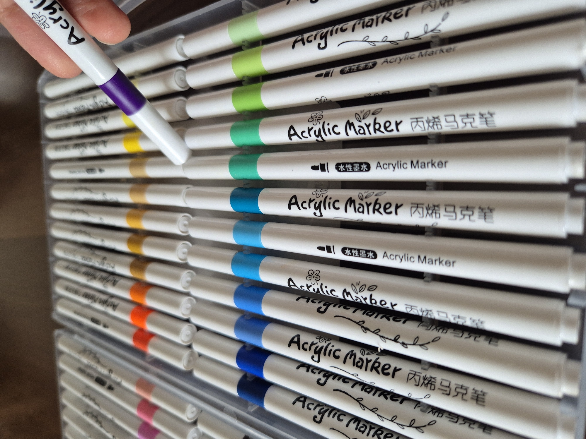 A hand is holding a white acrylic marker with a purple band near the tip, with the text "Acrylic Marker" and a floral design visible. The marker is positioned over a clear plastic case containing a set of acrylic markers, also white with colored bands near the tips, arranged in rows. The visible colors of the marker bands include purple, yellow, orange, brown, blue, cyan, green, and light green. The markers have black text labeling them as "Acrylic Marker" and Chinese characters. The background is a blurred wooden surface. There is no indication of Kokshetau, Kazakhstan in the image.