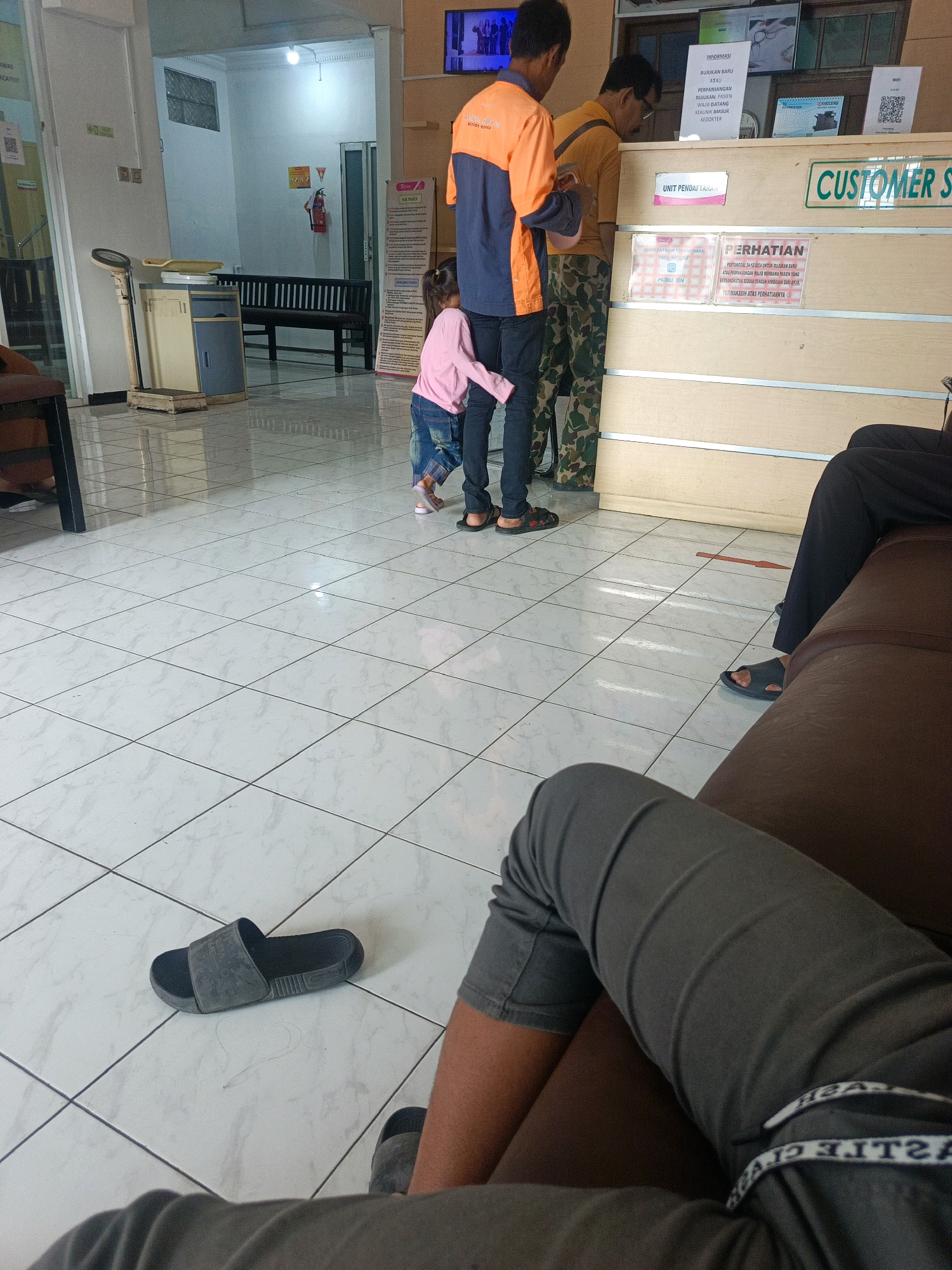 The image depicts the interior of a public facility, likely a government office or clinic, in Sedati Agung, Indonesia. The scene is brightly lit with fluorescent lighting and features tiled floors. In the foreground, a person is seated, their legs and lower torso visible, wearing grey pants and grey slides. To the left, a single black slide rests on the floor. In the mid-ground, two individuals stand behind a counter, one wearing an orange and blue uniform, and the other in a yellow shirt and camouflage pants. A young child in a pink shirt and blue jeans stands next to the person in uniform. Behind the counter, a sign reads "CUSTOMER SERVICE" in green lettering. Other signs and a calendar are visible on the counter. To the left of the counter, a medical scale and a cabinet are present, with a wooden bench and a fire extinguisher visible in the background. The overall environment suggests a functional waiting or service area.