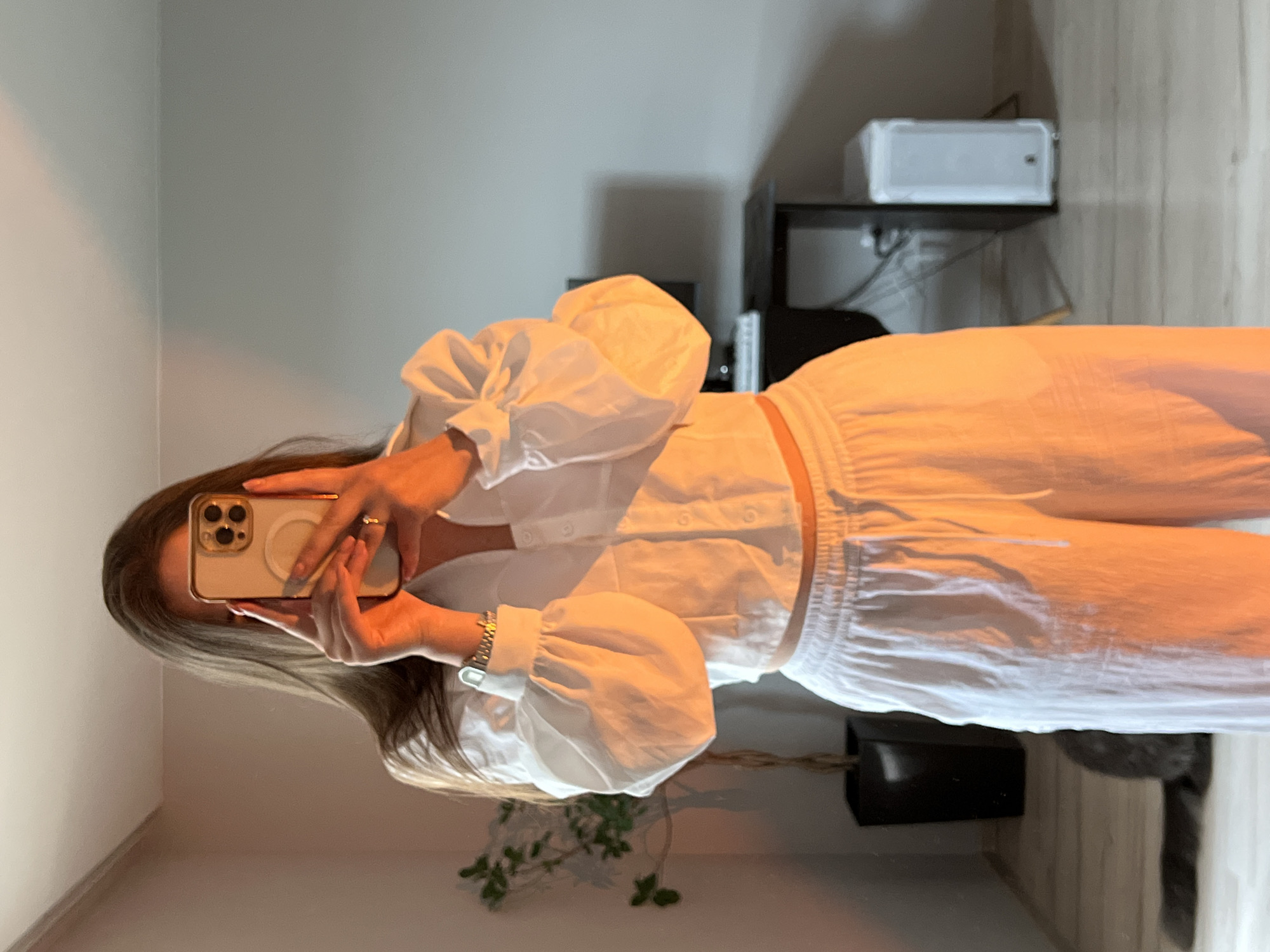 A mirror selfie taken indoors in Ternopil, Ukraine, captures a person (likely female) posing in a white outfit. The individual, with long brown hair, holds a light-colored smartphone with a triple camera array and a MagSafe accessory, effectively obscuring their face as they take the picture. They are dressed in a stylish white cropped button-up shirt featuring voluminous puffed sleeves, paired with matching white long pants that have an elasticated waist. Accessories include a gold-toned ring on their left hand and a silver-toned watch on their left wrist.
The reflection in the mirror reveals a room with white walls. In the background, a dark shelving unit holds a white electronic device, possibly a Wi-Fi router, along with other darker, less distinct objects. Cables and wires are visible behind the shelf. On the floor, a green-leafed plant in a dark pot is present, and a very blurry, dark shape in the lower right might be a pet. The warm, soft indoor lighting highlights the white clothing. No text is visible in the image.