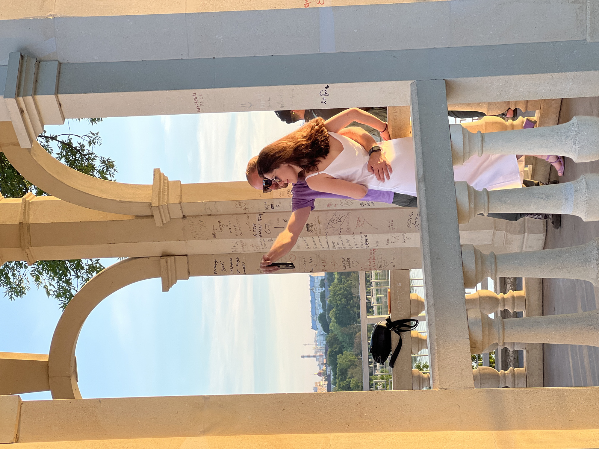 A man and a woman are taking a selfie in a sunlit, arched alcove. The man, wearing a purple t-shirt and sunglasses, has his arm around the woman, who is in a white dress and sunglasses. The alcove walls are covered in graffiti. In the background, a city skyline with trees and a body of water is visible. This is likely the Ternopil pond embankment in Ternopil, Ukraine.