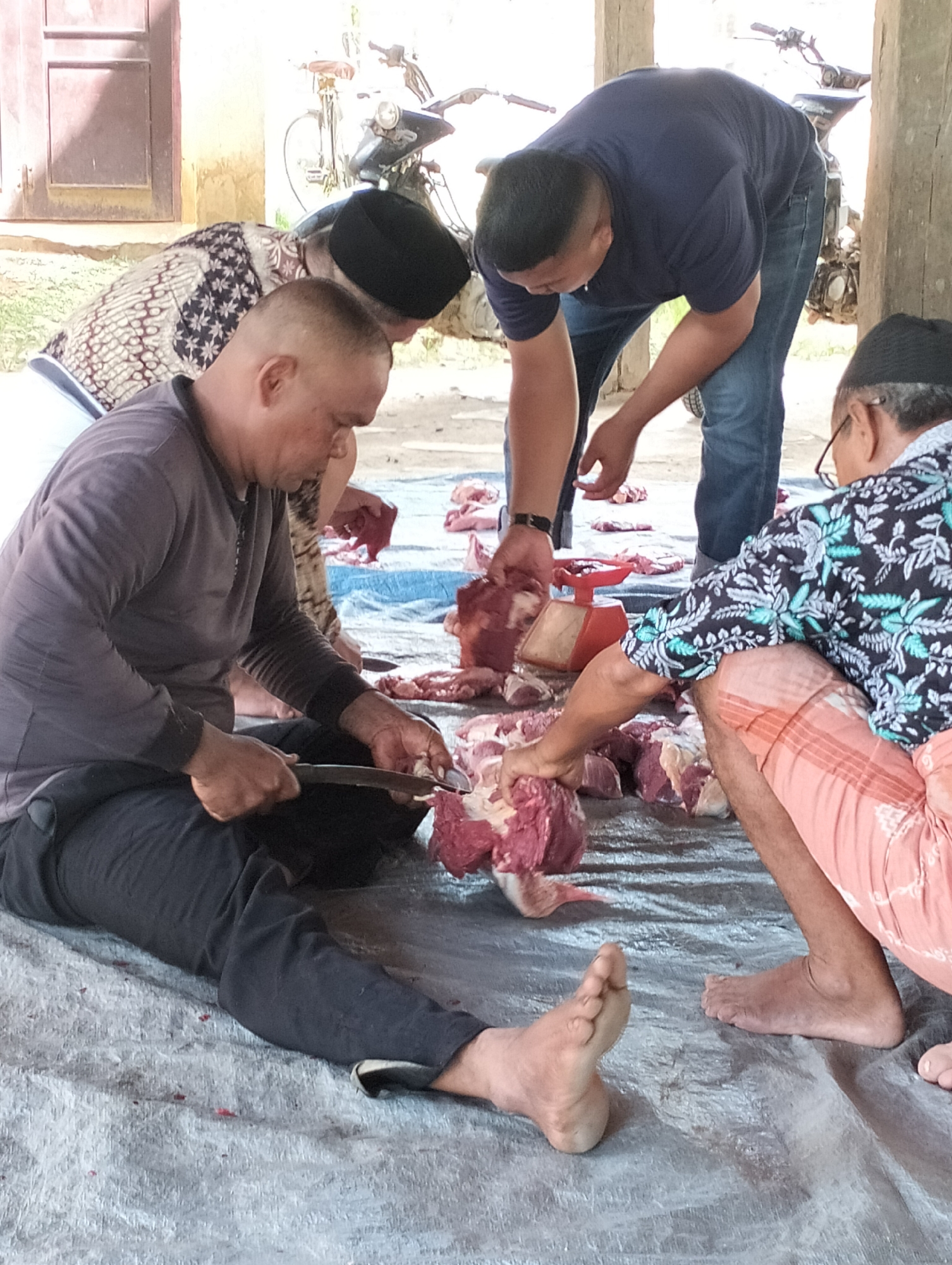 Four men are processing meat on a blue tarp outdoors in Meunasahalue, Indonesia. One man sits on the tarp wearing a long-sleeved gray shirt and dark pants, cutting meat with a knife. Another man, wearing a patterned batik shirt and a black skullcap, is handling meat near the seated man. A third man in a dark blue t-shirt and jeans is standing, also handling meat. The fourth man, wearing a dark floral shirt and pink patterned shorts, kneels on the tarp, handling meat near a small red scale. There are pieces of raw meat scattered across the tarp. A motorcycle and bicycle are in the background. The scene is outside a building with a reddish-brown door.