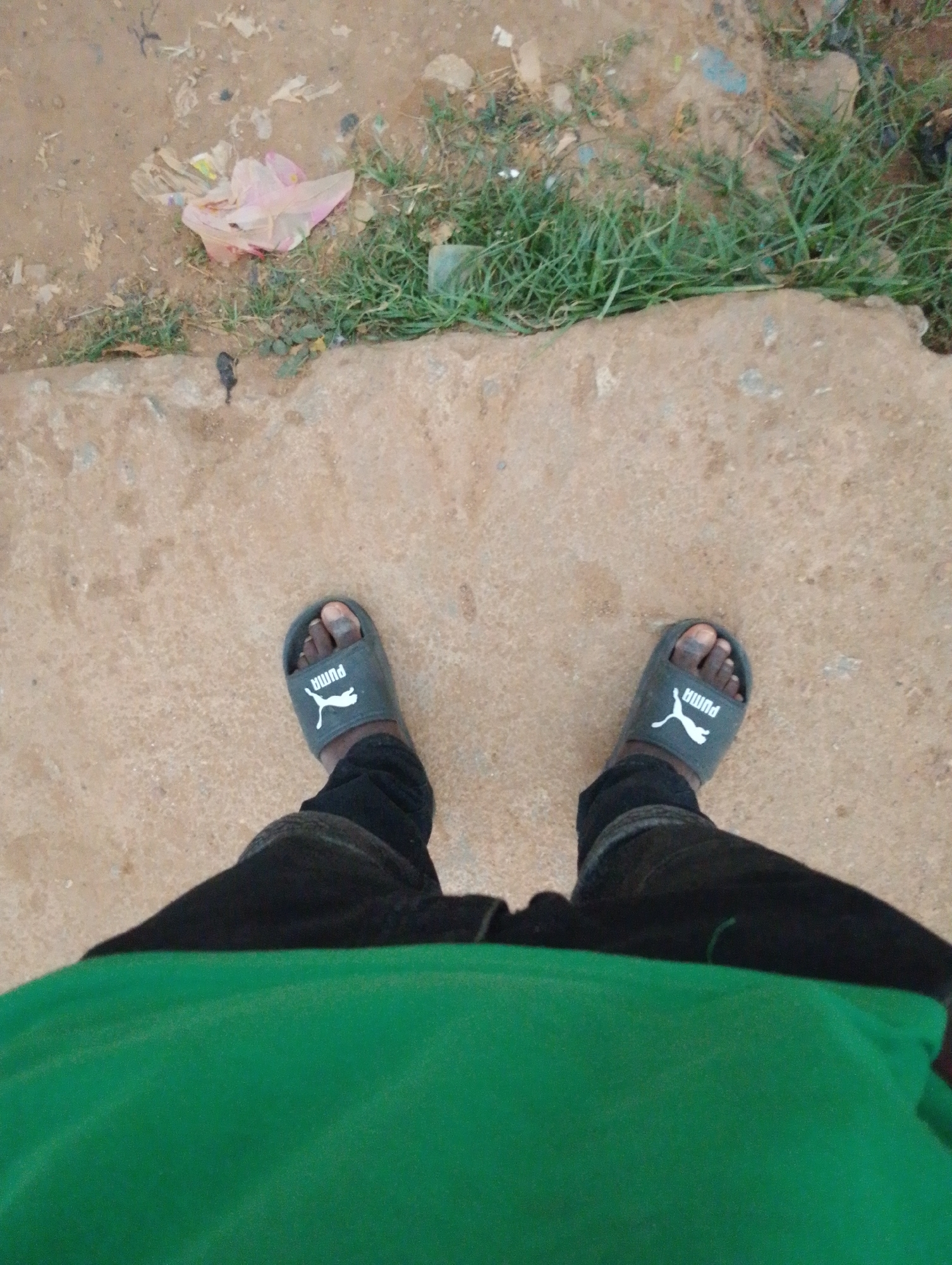 This is a first-person perspective image taken in Sabon Tasha, Nigeria. The scene depicts a person's feet wearing dark gray Puma slides and black pants. A bright green fabric, possibly clothing, is prominently featured in the foreground, obscuring the lower portion of the legs. The ground is a mix of dirt, sand, and a concrete path. Scattered on the dirt and grass area are pieces of litter, including a crumpled pink plastic bag. The lighting suggests it is daytime.