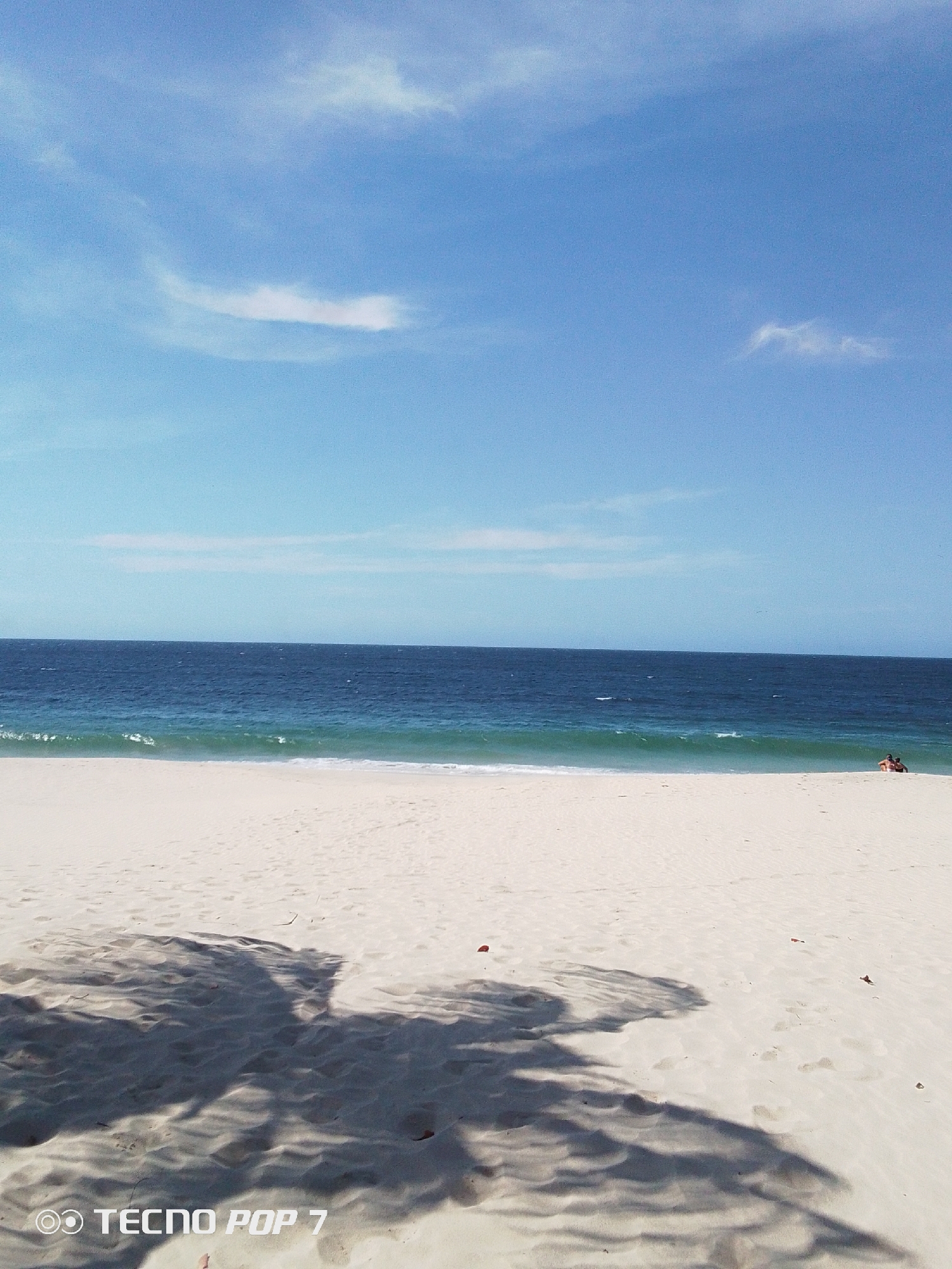The image depicts a beach scene in Pedro González, Venezuela. The sky is blue with scattered white clouds. The sea is dark blue, with lighter turquoise waves breaking on the white sand. A large shadow, likely from a tree, dominates the foreground of the beach. Two people are visible in the distance near the water's edge. The lower left corner shows the phone model "TECNO POP 7" imprinted on the image.