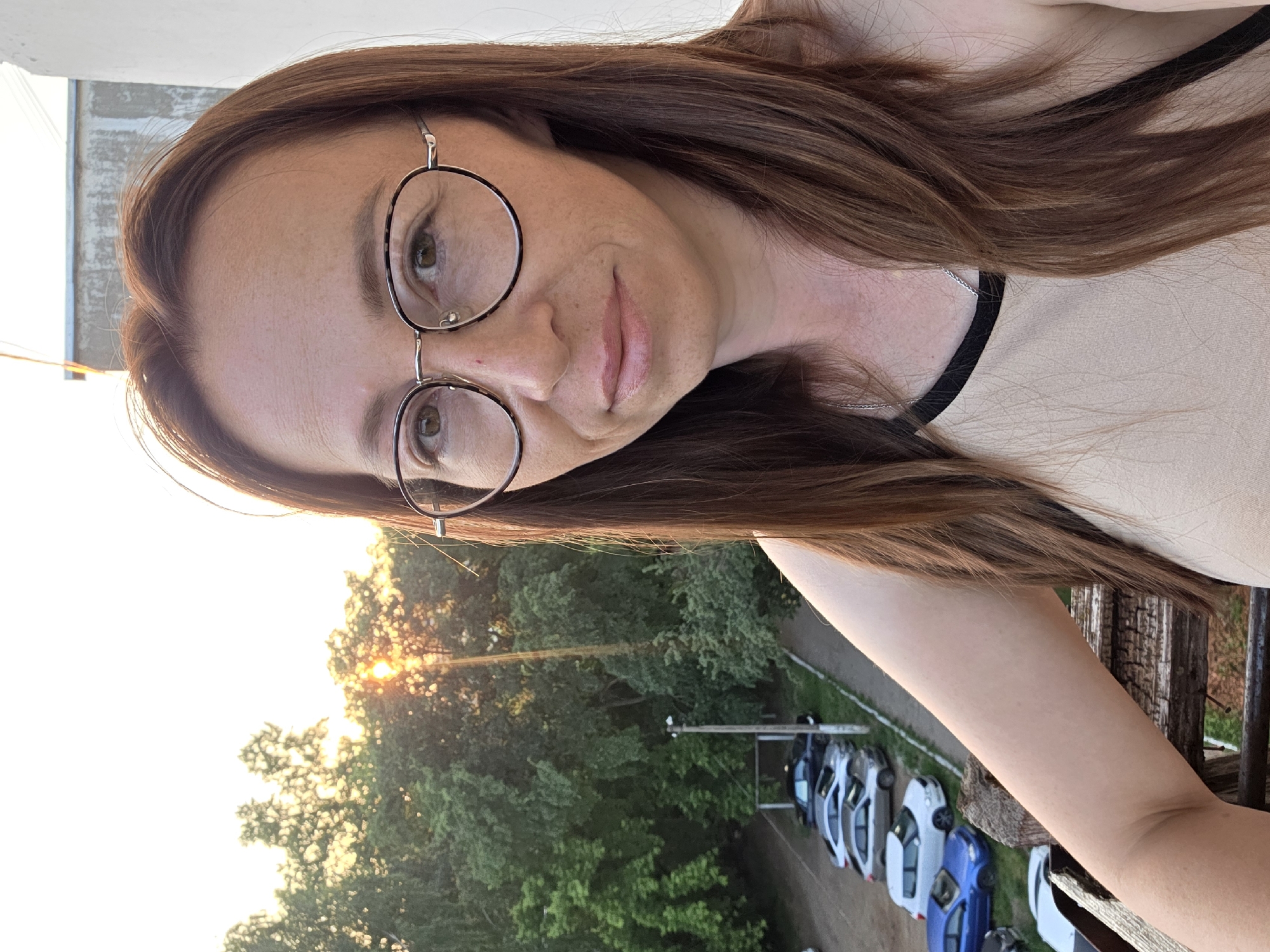 In Chaikovsky, Russia, at golden hour, a woman with long brown hair and glasses is taking a selfie on a balcony overlooking a street with parked cars and trees. The setting sun is visible behind the trees, casting a warm glow. The woman is wearing a light-colored top with a dark trim. The balcony railing is made of weathered wood.