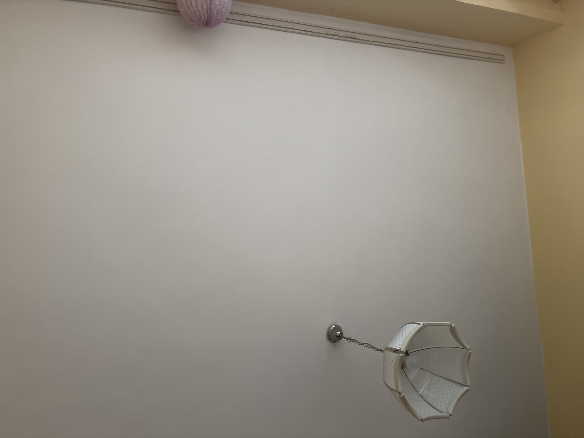 An indoor shot captures a section of a ceiling and upper wall. A white, octagonal lampshade hangs from a silver chain attached to the ceiling. To the right, a portion of a light purple, spherical decorative item is visible, along with a vertical channel molding on the wall. The ceiling is painted a neutral white, and the upper part of the wall is a pale yellow. No people or specific locations within Sofia, Bulgaria, are identifiable.
