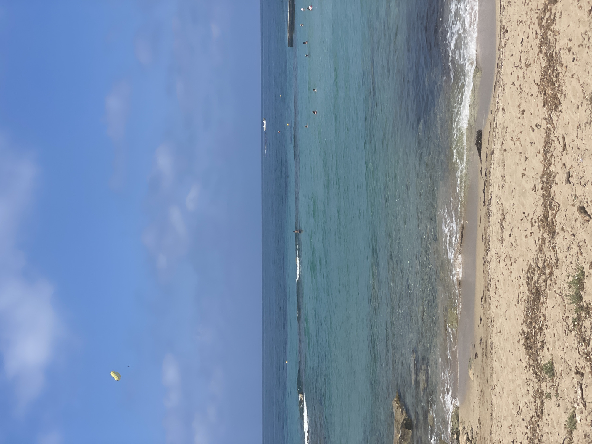 The image depicts a beach scene on a clear day in Paphos Municipality, Cyprus. The foreground shows a sandy beach with scattered dried vegetation. The sea is a clear turquoise color, with gentle waves lapping at the shore. Several people are visible in the water, swimming and enjoying the sea. Further out, a boat can be seen moving across the water, and a parasail is visible in the sky. The horizon is lined with a darker blue sea, meeting a bright blue sky dotted with white clouds. A concrete pier or breakwater extends into the sea on the right side of the image. The overall impression is one of a peaceful and idyllic coastal setting.
