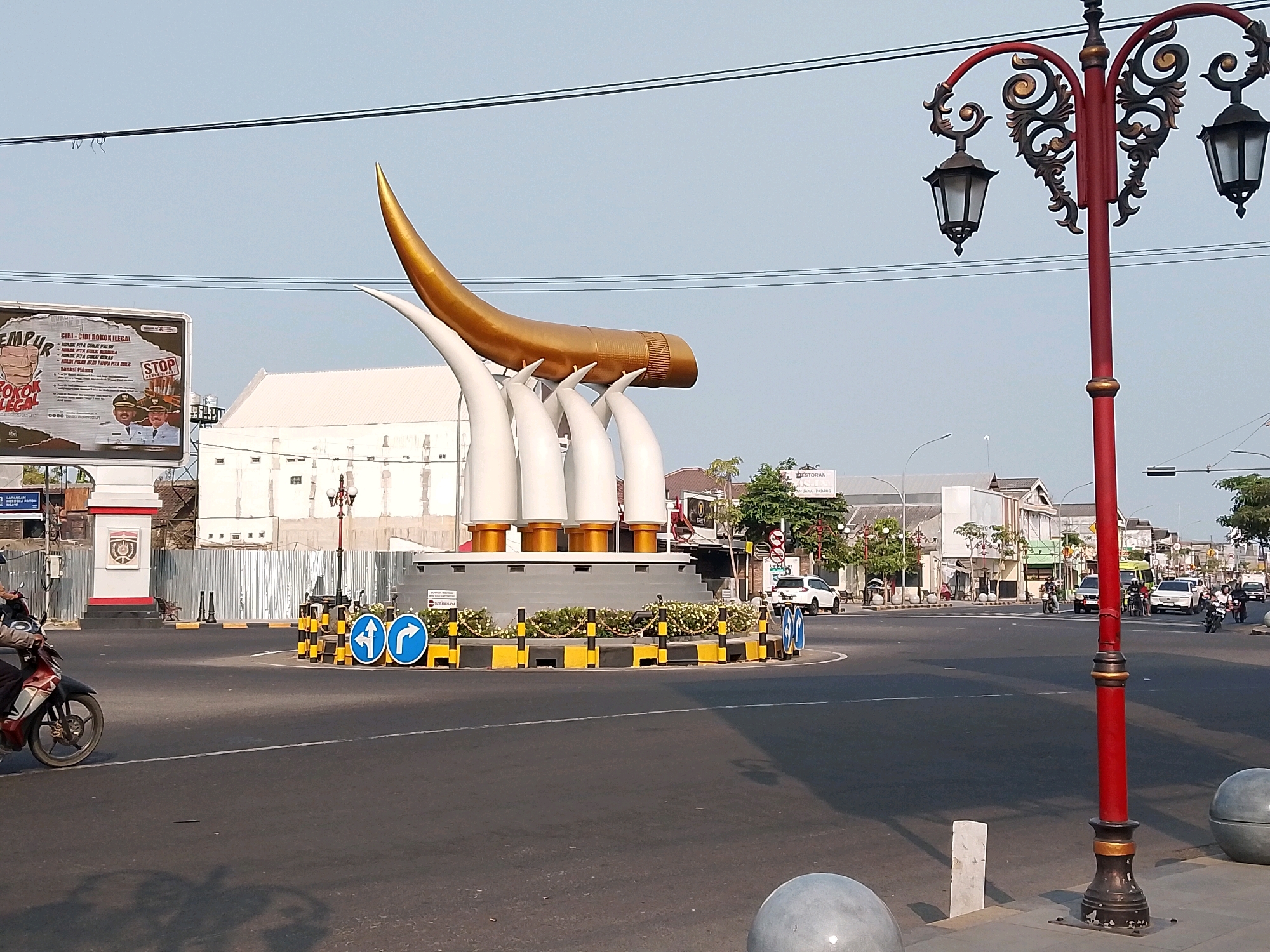 This image captures a street scene in Ngawi, Indonesia, with a prominent roundabout featuring a golden horn-shaped sculpture at its center. The sculpture is supported by white, horn-like structures. Around the roundabout, traffic signs indicate directional arrows and a prohibition to enter. Decorative bollards with yellow and black stripes line the roundabout's edge, interspersed with small white flowers.
To the left of the roundabout, a billboard displays information related to "SMOKING LEGAL" with images of two men in uniform and a "STOP" sign. Adjacent to the billboard is a white building with a red and white arched entrance and a crest. A construction fence partially obscures the background.
On the road, a person is riding a red and white motorcycle in the foreground, casting a shadow on the asphalt. Various vehicles, including cars and motorcycles, are visible in the background, navigating the intersection. A decorative red lamppost with ornate black detailing and three lamps stands prominently in the right foreground. The environment is characterized by clear sky, asphalt roads, and several buildings lining the street, suggesting an urban setting.