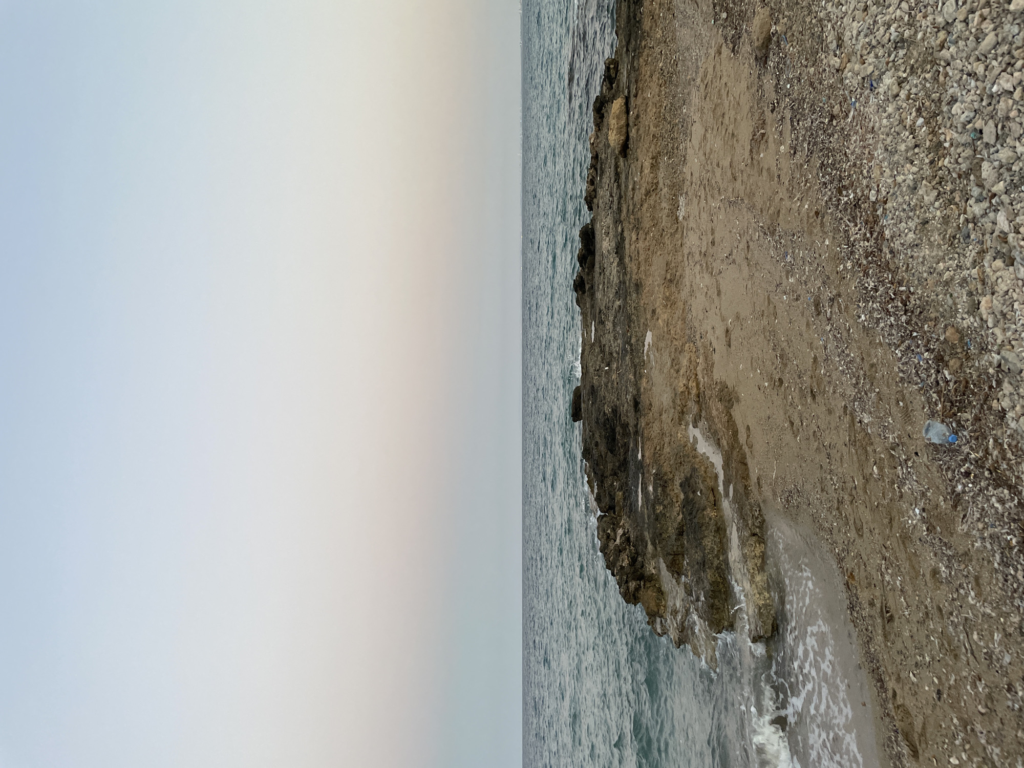 A wide-angle, eye-level shot captures a tranquil seaside scene at dusk. The sky, a gradient of pale blue and soft pink, meets the dark, rippling ocean horizon. In the foreground, rough, dark rocks emerge from the water, creating a jagged coastline. The shore transitions from wet sand at the water's edge to drier, coarser sand mixed with small pebbles. A single, clear plastic water bottle with a blue cap lies on the pebbled area to the right. The overall impression is one of natural beauty and peaceful solitude. This location is identified as Trikomo, Northern Cyprus / Cyprus.