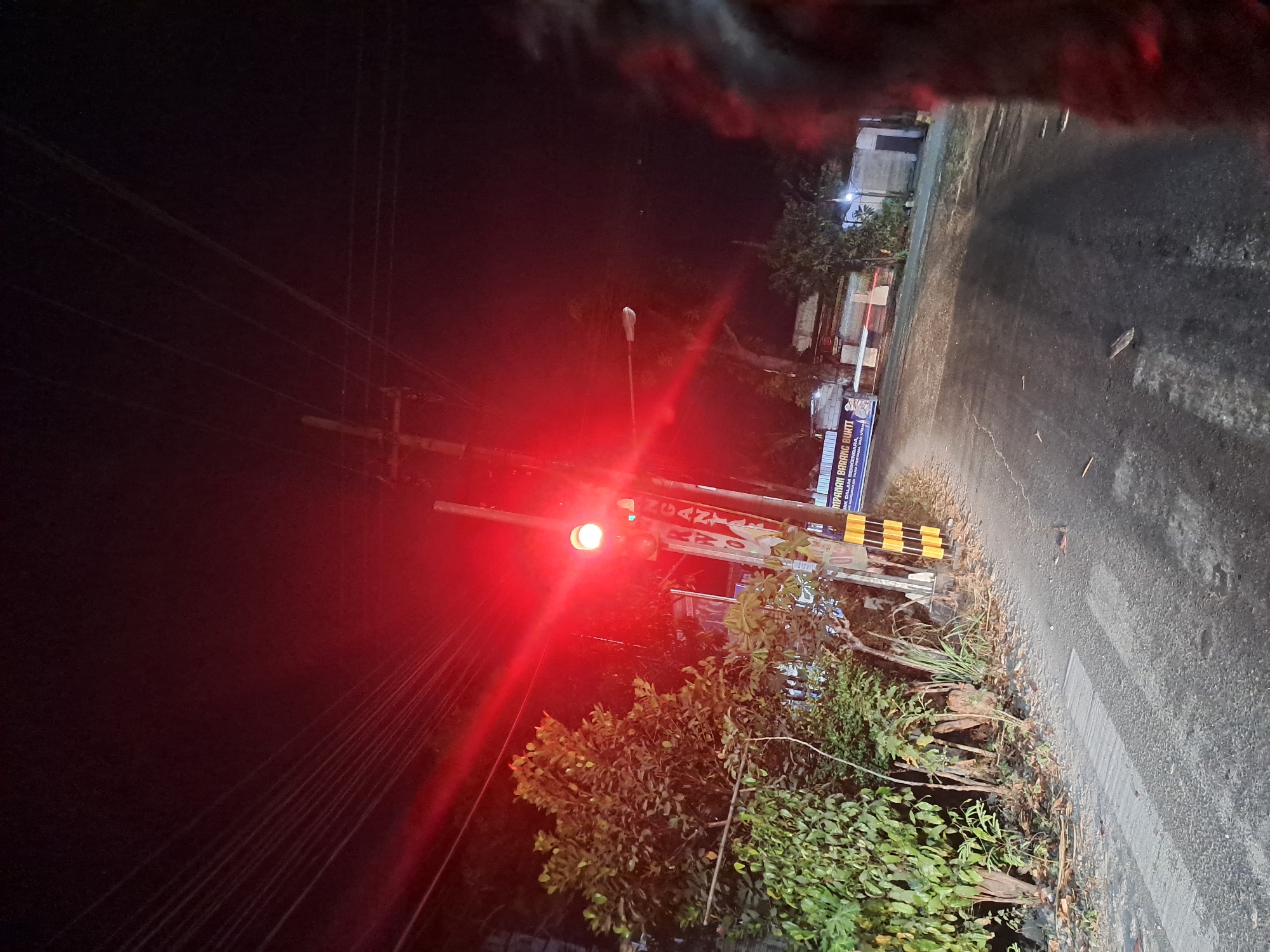 This is a nighttime photograph taken at an outdoor location, likely a roadside in Slambur, Indonesia, as indicated by the presence of a red traffic signal and local signage. The image is dominated by the bright red glow of a traffic light, which casts a strong illumination on the surrounding area, creating lens flares and washing out details in the upper portion of the frame.
In the foreground, a paved road curves to the right. To the left of the road, there is lush green vegetation, including trees and bushes. A utility pole with a traffic light is positioned on the left side of the road, with the red light illuminated. The pole itself has yellow and black striped markings near its base, typical of warning or safety markers. There is also some signage on or near the pole. Further down the road, some buildings and additional streetlights are visible in the background, illuminated by the ambient light and the traffic signal. The sky is dark, indicating nighttime. The overall impression is of a quiet street at night, with the traffic light signaling a stop.