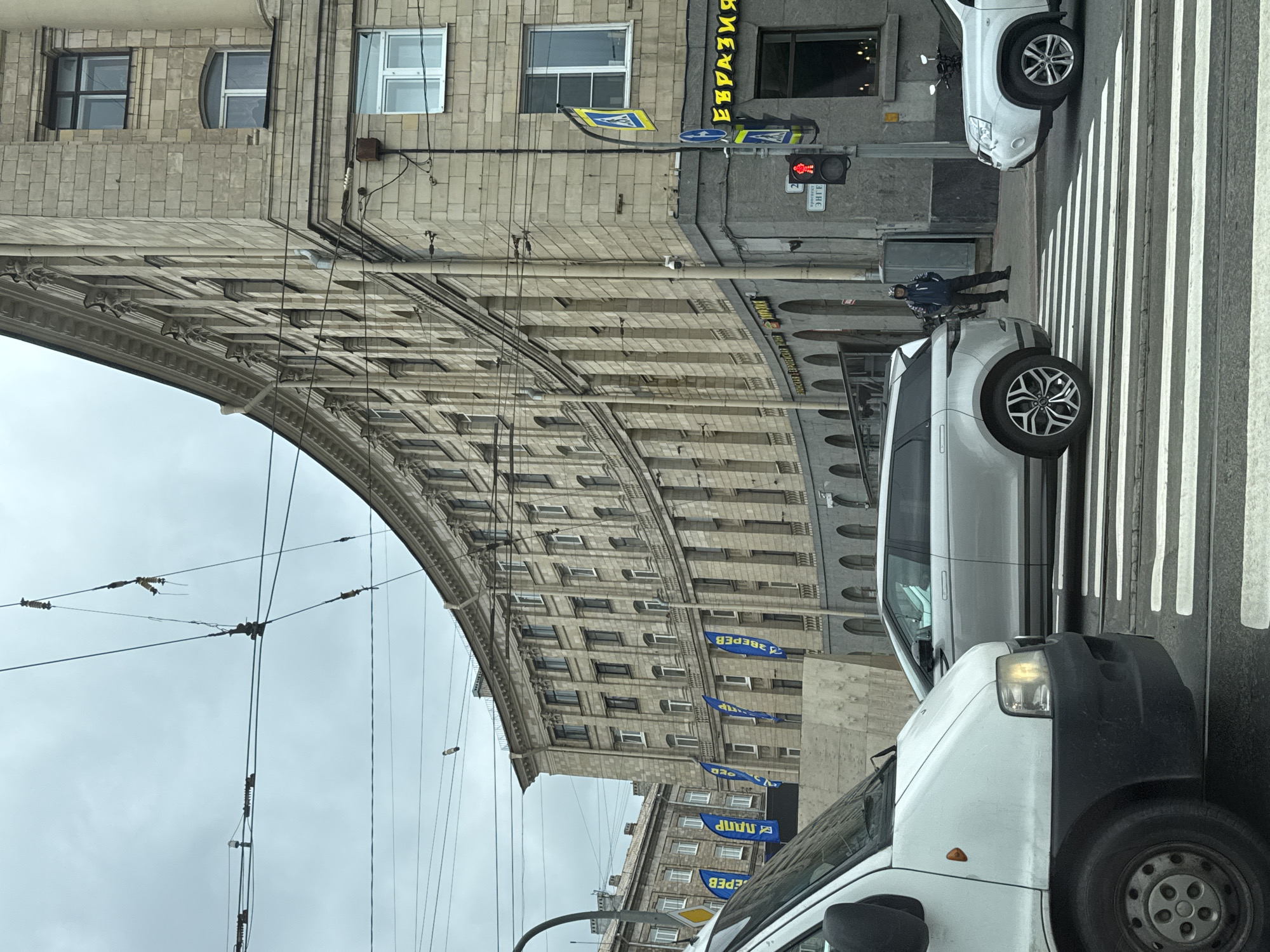 This image captures a street scene in Saint Petersburg, Russia, specifically near the Singer House building on Nevsky Prospekt. The perspective is from a vehicle at a crosswalk, looking towards the curved facade of a large, stone building. The sky is overcast.
In the foreground, the front of a white van is visible on the left, and a silver SUV is crossing the crosswalk in the middle of the frame. On the right, a white SUV is stopped at the crosswalk. A pedestrian wearing dark clothing and a helmet stands on the sidewalk near the entrance to the building.
The building itself is a multi-story structure with a distinct curved architecture, clad in light-colored stone blocks. It features numerous windows, some with dark frames and others with white frames. Decorative architectural elements, such as cornices and pilasters, are visible along the facade. Several blue flags with white lettering are displayed along the lower part of the building. Above the street, a complex network of trolley wires is visible against the sky.
Street signs are present, including a pedestrian crossing sign with a yellow background and a blue circular sign with a white arrow. A traffic light shows a red pedestrian figure. A sign with yellow Cyrillic lettering, reading "ЕВРАЗИЯ" (Eurasia), is visible above a storefront. Another smaller sign below it has Cyrillic lettering that appears to read "КРАСКИ ЭНГЕЛЬ" (Paints Engel). A street number "2" is also visible.
The scene depicts traffic and pedestrian activity at an intersection under cloudy weather conditions.