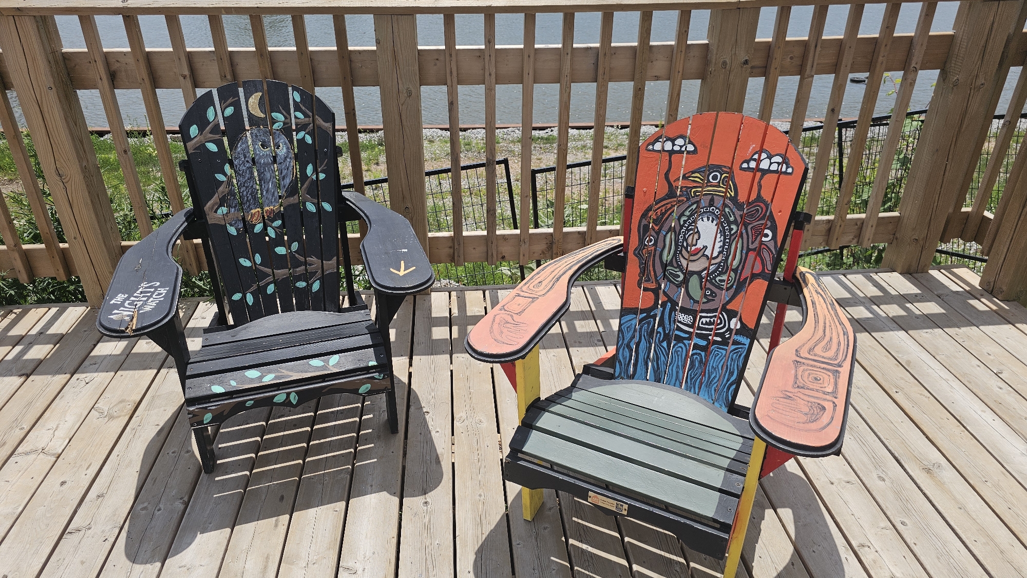 Two Adirondack chairs are positioned on a wooden deck. The chair on the left is black and features a painted owl on a branch with a crescent moon above. The text "THE NIGHT'S WATCH" is visible on its armrest. The chair on the right is primarily orange and features a vibrant, colorful painting of a figure with an elaborate headdress. The deck is made of light-colored wooden planks. Behind the chairs, a wooden railing overlooks a body of water. The background shows greenery and a fence line. The lighting suggests it is daytime, possibly late morning or early afternoon, with distinct shadows cast by the chairs. The scene appears to be a serene outdoor setting, likely a patio or deck area near a waterfront, given the context of Oakville, Canada. There is no activity taking place; the chairs are empty.