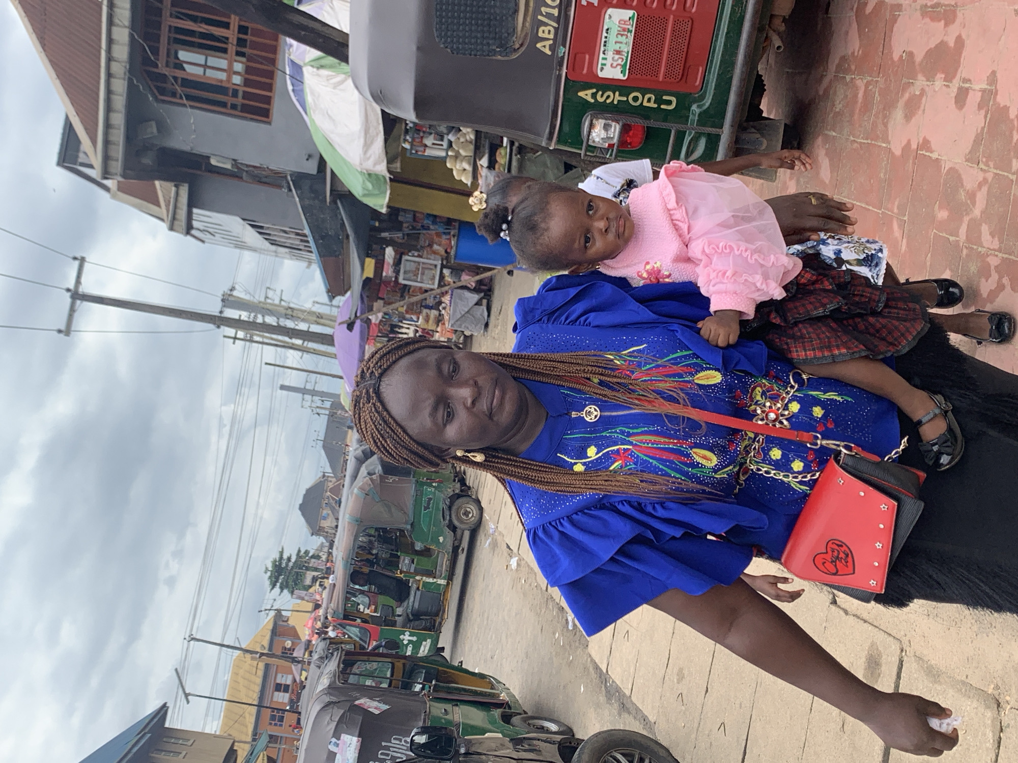In Umuode, Nigeria, a woman with braided hair carries a baby dressed in a pink ruffled top and black shoes. The woman wears a blue dress adorned with colorful patterns and rhinestones, accessorized with a gold necklace and a red crossbody bag. She is walking on a paved area, with several auto-rickshaws visible in the background. The environment appears to be a street market or busy thoroughfare, with buildings and market stalls lining the sides. The sky is overcast.