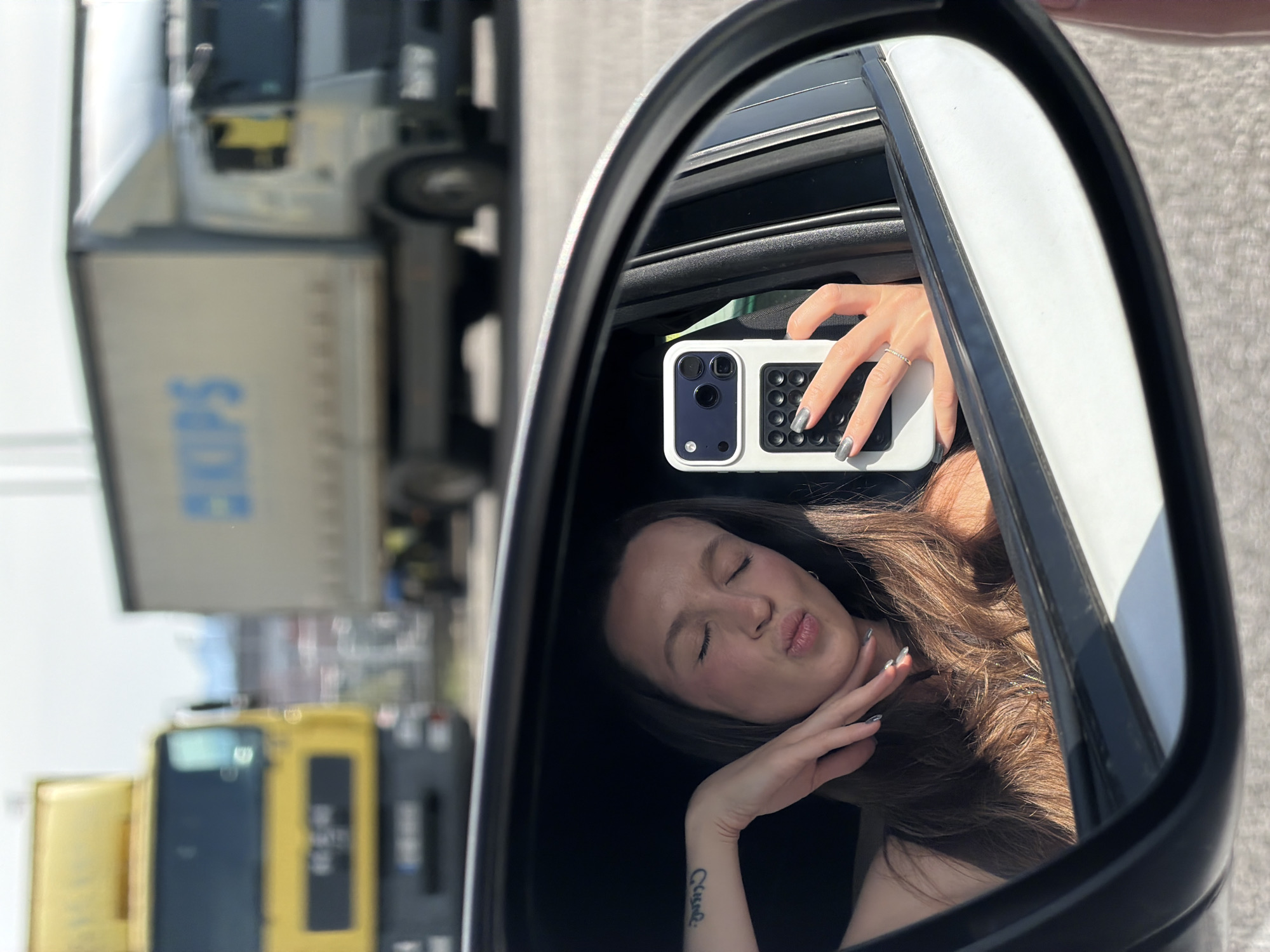A young woman is visible in a car side mirror, presumably taking a selfie. She has long brown hair and is puckering her lips with her eyes closed. Her left hand is near her face, and a tattoo is visible on her left wrist. She holds a white phone in her right hand. The reflection shows the interior of the car as well as blurred trucks on a road. The word "SKIPS" is visible on one of the trucks. The location is Donja Cijevna, Montenegro.