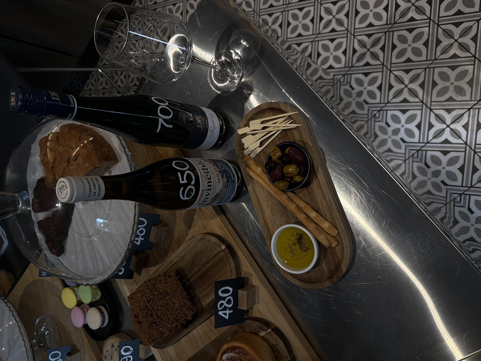 The image presents a high-angle shot of a spread of food and drink on a metallic countertop with a patterned background. The scene includes two wine bottles labeled "700" and "650," a clear wine glass, and several wooden platters arranged with assorted items.
On one platter, there are olives, breadsticks, and a small bowl containing a yellow oil with black specks. Another platter showcases macarons in pastel colors and a slice of dense bread. There are small price tags with numbers such as "400" and "480" next to each platter.
The background features a black and white geometric tile pattern.
Given the scene's characteristics—wine, olives, and other delicacies—it is likely that the location is a restaurant in the city of Belgrade, Serbia.