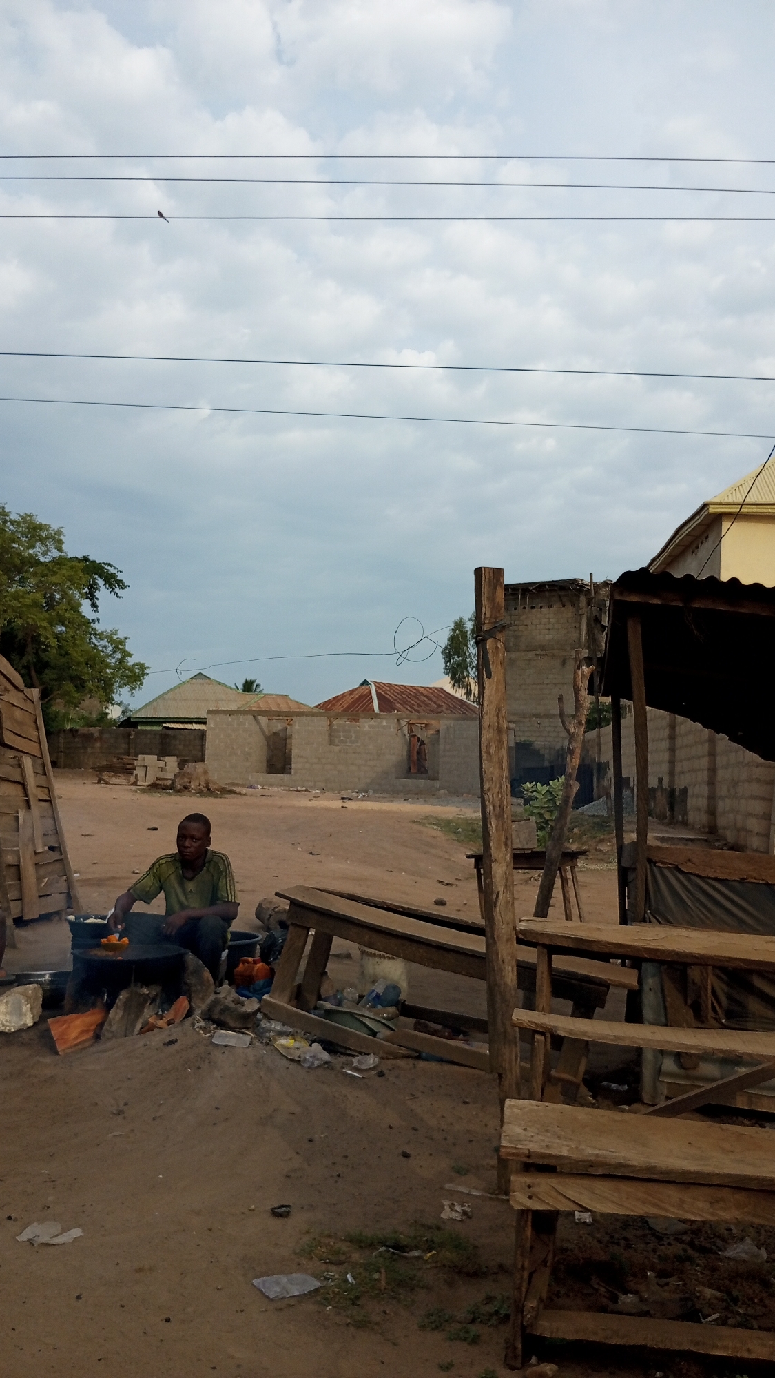 This image depicts an outdoor scene in Ajaokuta, Nigeria, under an overcast sky. In the foreground, a man in a green shirt and dark trousers sits on the dirt ground, actively cooking. He is tending to a pot or pan placed over a makeshift stove or open fire, with bright orange food items, likely fried plantains or yams, visible inside. Nearby, a black bucket and various discarded items, including plastic bottles and wrappers, are scattered on the unpaved ground.
To the right, a series of rustic wooden benches and a simple stall-like structure with a dark, perhaps corrugated iron, roof are prominent. A sturdy wooden post stands upright in the mid-ground. The entire ground area is unpaved, consisting of reddish-brown dirt.
In the background, several buildings are visible. Some appear to be under construction with exposed cinder block walls, while others have finished structures with corrugated metal roofs, some showing signs of rust. Green trees and foliage are visible on the left side of the frame. Overhead, multiple black power lines stretch across the cloudy, light-gray sky, with a small dark bird perched on one of the higher lines. The soft, diffuse lighting suggests an overcast day or possibly early morning/late afternoon. There is no visible text in the image. The scene captures an everyday moment of outdoor cooking, likely street food vending, in a semi-urban or rural Nigerian setting.