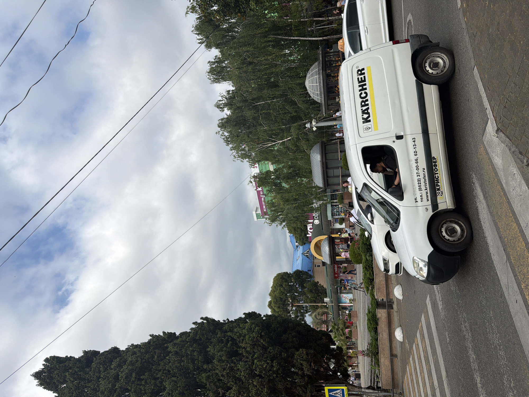 This media file captures a street scene in Sochi, Russia, on an overcast day. The primary focus is a white Kärcher branded van parked on the right side of the road, oriented sideways in the frame. The van's side displays the Kärcher logo in yellow and black, along with contact information including a street name, phone numbers, and a website address: "ул. Орджоникидзе, 8 тел.: (8622) 37-00-66, 62-13-43 www.kristofer.ru КРИСТОФЕР". A person is visible inside the van, seated in the driver's seat.
In the background, a bustling street is lined with various buildings, some featuring storefronts with signage. Notable elements include a building with a bright blue roof and a distinctive domed structure. Lush green trees, including a large, dense tree in the foreground on the left, add to the urban greenery. Overhead, power lines crisscross the sky, which is filled with grey, cloudy masses.
To the left of the van, another white car is partially visible, also parked on the street. A pedestrian crossing is marked on the asphalt in front of the van. People can be seen walking along the sidewalk and in the street, suggesting a typical day in a commercial or residential area. The overall impression is of an ordinary day with urban activity under a cloudy sky.