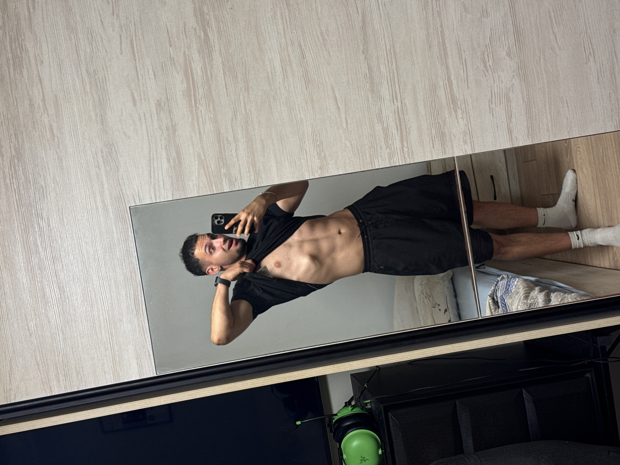 A young man with short dark hair and a beard is standing in front of a mirror, taking a selfie with his phone. He is wearing a black t-shirt, which he has pulled up to reveal his abdominal muscles, and black shorts. He is also wearing white socks. The mirror reflects his body from the waist up and also shows a bed with a patterned duvet behind him. To the left of the mirror is a tall, dark cabinet or wardrobe. On top of this cabinet, to the left of the mirror, is a pair of bright green headphones with a black microphone. The floor appears to be made of light-colored wood. The background wall has a light wood-grain texture. The scene appears to be indoors, likely in a bedroom or a dressing area. There is no indication of the city of Shumen, Bulgaria, in the image.