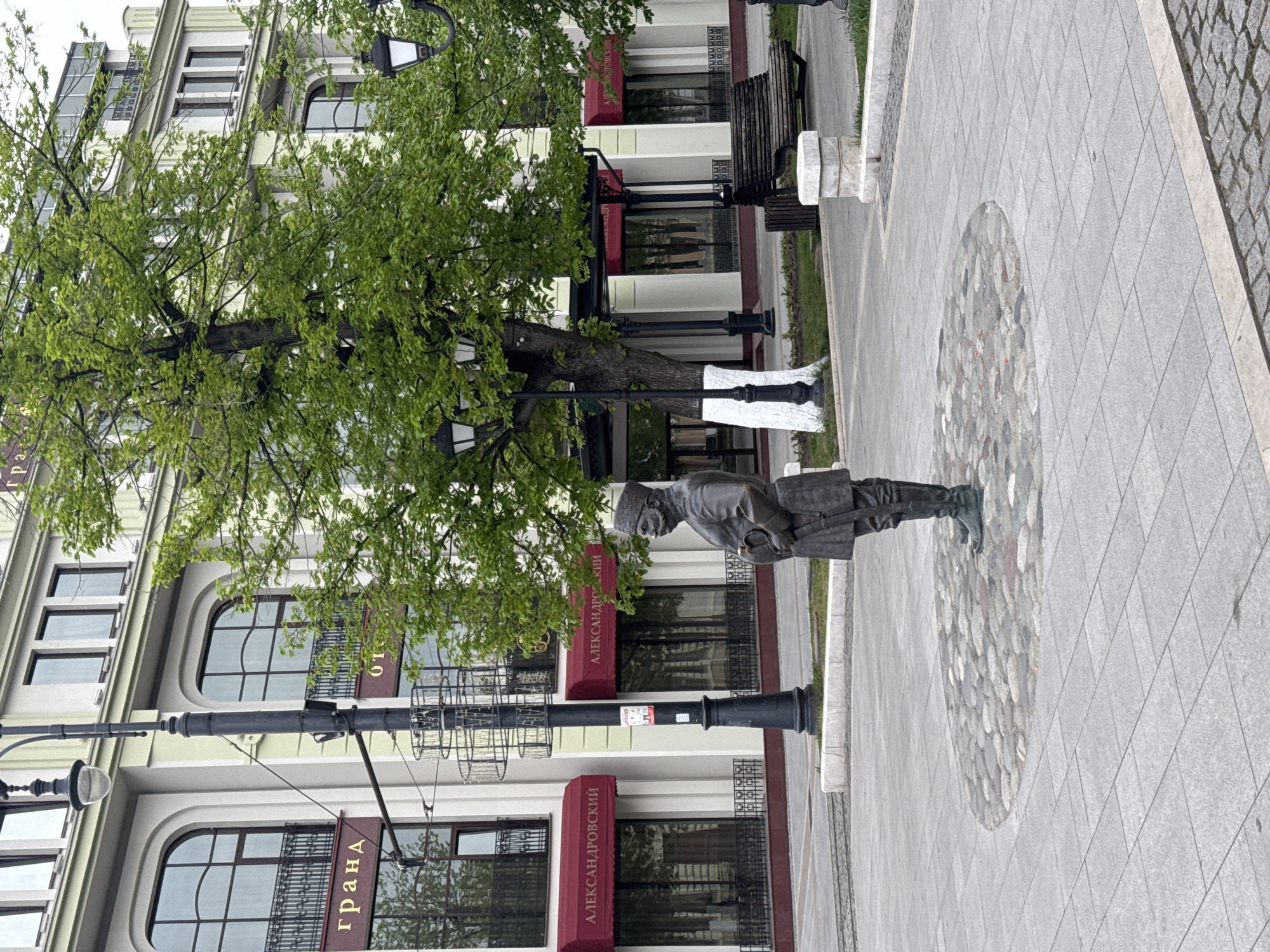 The image shows a statue of a man standing on a cobblestone mosaic embedded in a paved sidewalk. Behind the statue is a multi-story building with signs in Cyrillic script, indicating a location in a region with Russian-speaking influence. The building has dark-framed windows with metal railings and dark red awnings over the windows. A tree grows near the statue and partially obscures the view of the building. The image appears to be taken in Akhieli, Georgia, based on the Cyrillic signage.