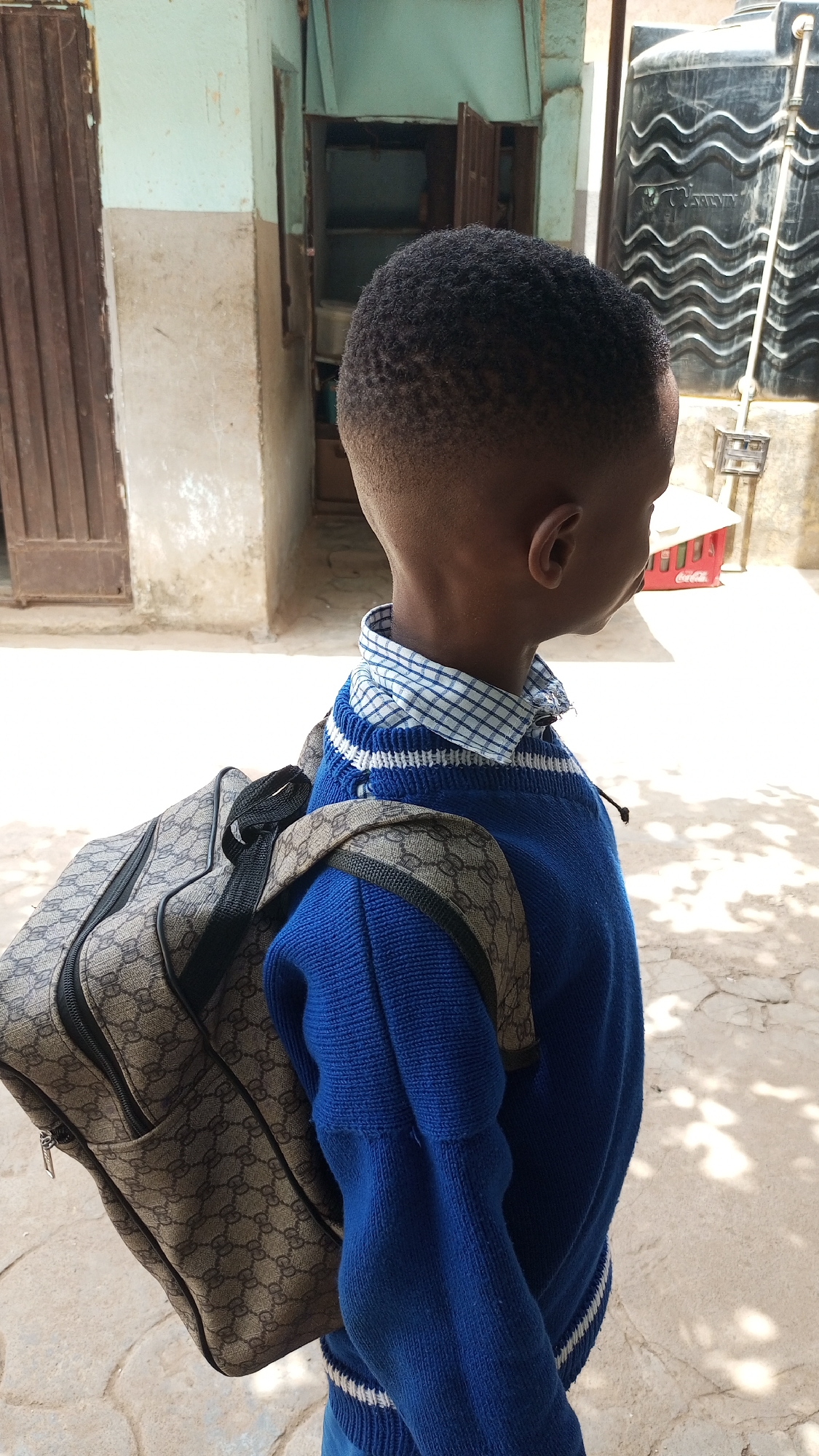 A young boy in Sabon Tasha, Nigeria, is seen from the side, wearing a blue sweater over a checkered shirt and carrying a patterned backpack. He stands on a paved surface, possibly a sidewalk, with buildings and a water tank visible in the background. A brown door is to the left, with another building structure behind. A Coca-Cola crate is near the building on the right. The area is shaded by trees.