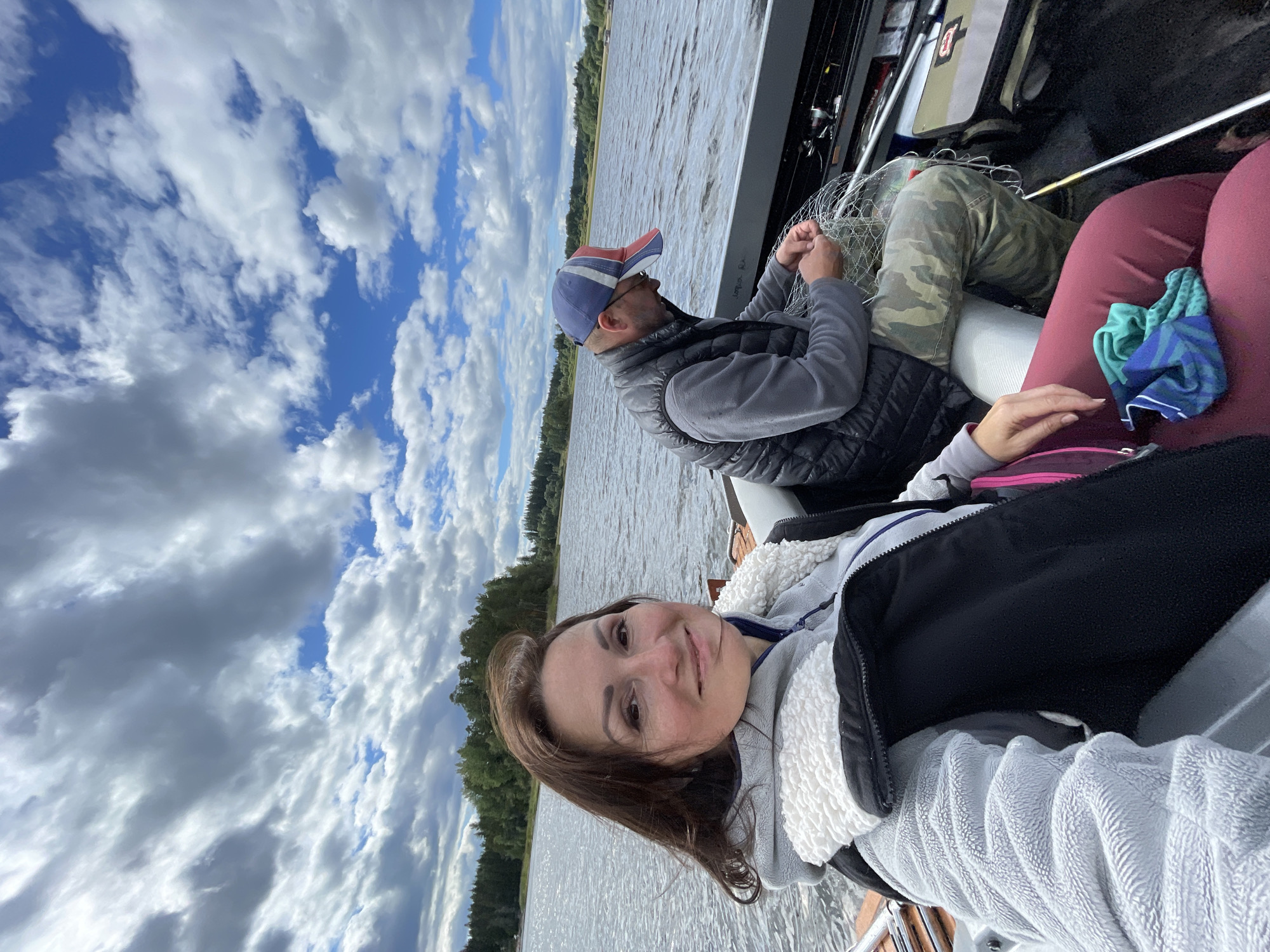 A woman and a man are in a boat on a body of water. The woman is in the foreground, looking at the camera and smiling. She has long brown hair and is wearing a grey fleece jacket with a white fluffy collar and a black vest. The man is in the background, wearing a grey long-sleeved shirt, a dark puffy vest, camouflage pants, and a red, white, and blue baseball cap. He is focused on untangling a fishing net. The water is choppy, and the sky is cloudy with patches of blue. Trees and land are visible in the distance. The location is the Beryozovo district in Russia, where Beryozovo is a town.