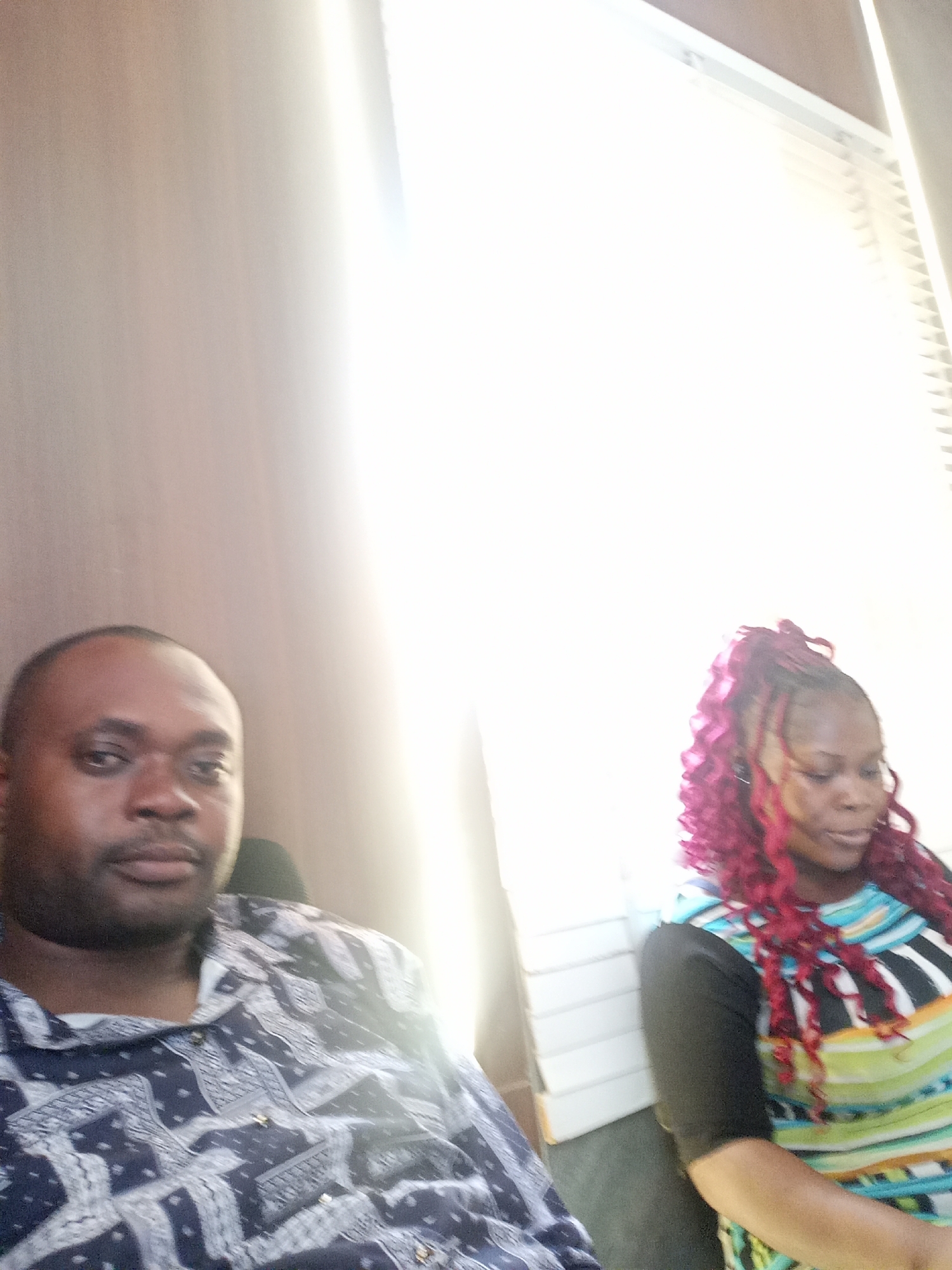 Two people are in the frame. A dark-skinned man with a shaved head is on the left. He is wearing a dark blue patterned shirt. A dark-skinned woman is on the right. She has red hair in a curly style and is wearing a black-sleeved shirt with a horizontally striped bodice. The background is a wall and a window with white blinds. The location cannot be verified to be Gwarinpa, Nigeria, from this image.
