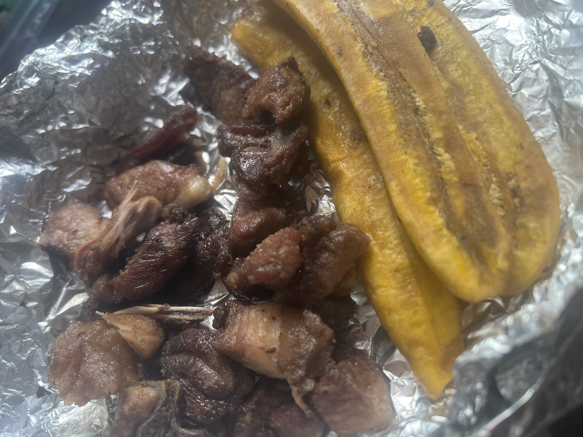 This image shows a meal likely purchased and consumed in Dar es-Salaam, Tanzania. The meal consists of fried plantains (likely called 'ndizi' locally) and fried meat, likely goat or beef, served on a sheet of aluminum foil. The foil suggests it may be a takeaway meal from a local vendor or restaurant, a common food service practice in Tanzania. The plantains are ripe, evidenced by their yellow color and some dark patches. The meat appears to be cut into small pieces, possibly seasoned, and fried until browned. The meal is a typical street food offering in Dar es-Salaam.
