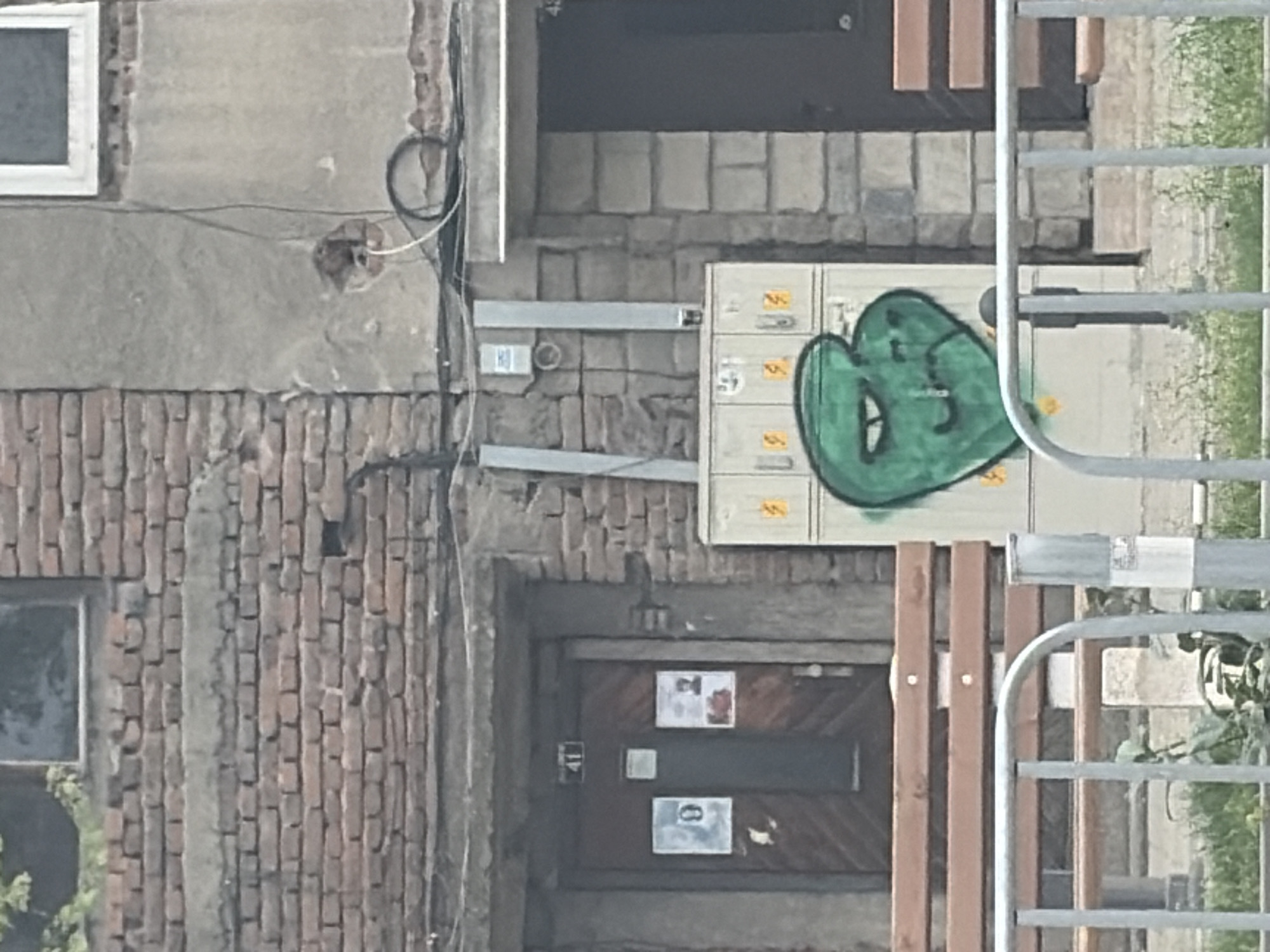 The image depicts a fragment of an urban street scene, likely in Plovdiv, Bulgaria, featuring building facades, utility infrastructure, and a metal fence. The original image is rotated 90 degrees counter-clockwise; described here as if upright.
On the left, a building wall is visible, transitioning from a lower section of weathered red brickwork to an upper section of lighter, worn plaster. A small, square window with a white frame is situated in the upper-left of the plastered wall. Black and white electrical wires are routed along this wall, entering through a small, rough hole.
In the central part of the image, an off-white or beige electrical utility box with multiple compartments is mounted on a wall constructed of light-colored, rough-cut stone blocks. A prominent piece of green graffiti, stylized as a cartoonish, blob-like face with large eyes and a simple mouth, is spray-painted on the right side of this box. Several small yellow stickers with numbers or symbols, such as "5" and "S", are affixed to the box's compartments. Below the box, two horizontal gray conduits or cable trays run along the stone wall.
To the right, a silver-colored metal fence or gate with a grid pattern partially frames the scene, revealing some green vegetation beyond.
At the bottom of the image, below the brickwork, a dark wooden door with diagonal paneling is visible. On this door, a white sign with the number "41" is vertically oriented, and another white sign below it displays the number "0". Other smaller notices are also attached to the door but are illegible. To the right of the door, several vertical brown wooden slats suggest another part of a fence or barrier.
The scene captures a typical daytime urban environment, characterized by functional infrastructure and a blend of old and practical elements.
