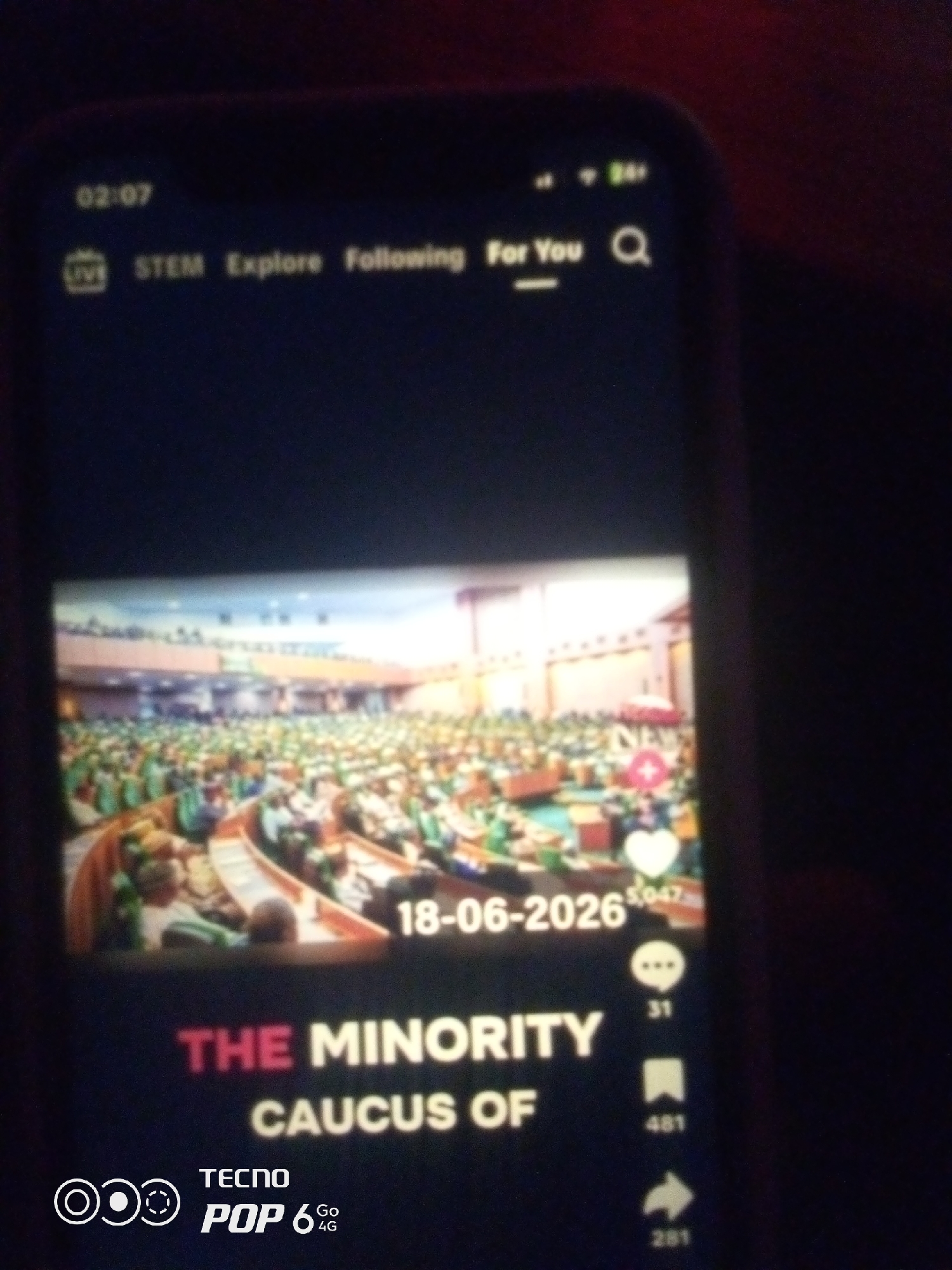 The image shows a mobile phone screen displaying a video. The video appears to be a recording of a large assembly, possibly a parliamentary session or a conference, with many people seated in an auditorium. The text on the screen reads "THE MINORITY CAUCUS OF" and a date "18-06-2026". There are also icons indicating engagement metrics like likes, comments, shares, and saves, with numbers associated with them. The lower part of the screen shows "TECNO POP 6 4G" branding. The phone is being held in a dark environment, with the background suggesting a dimly lit room. There is no visible indication of the city of Howayi, Nigeria in the image.