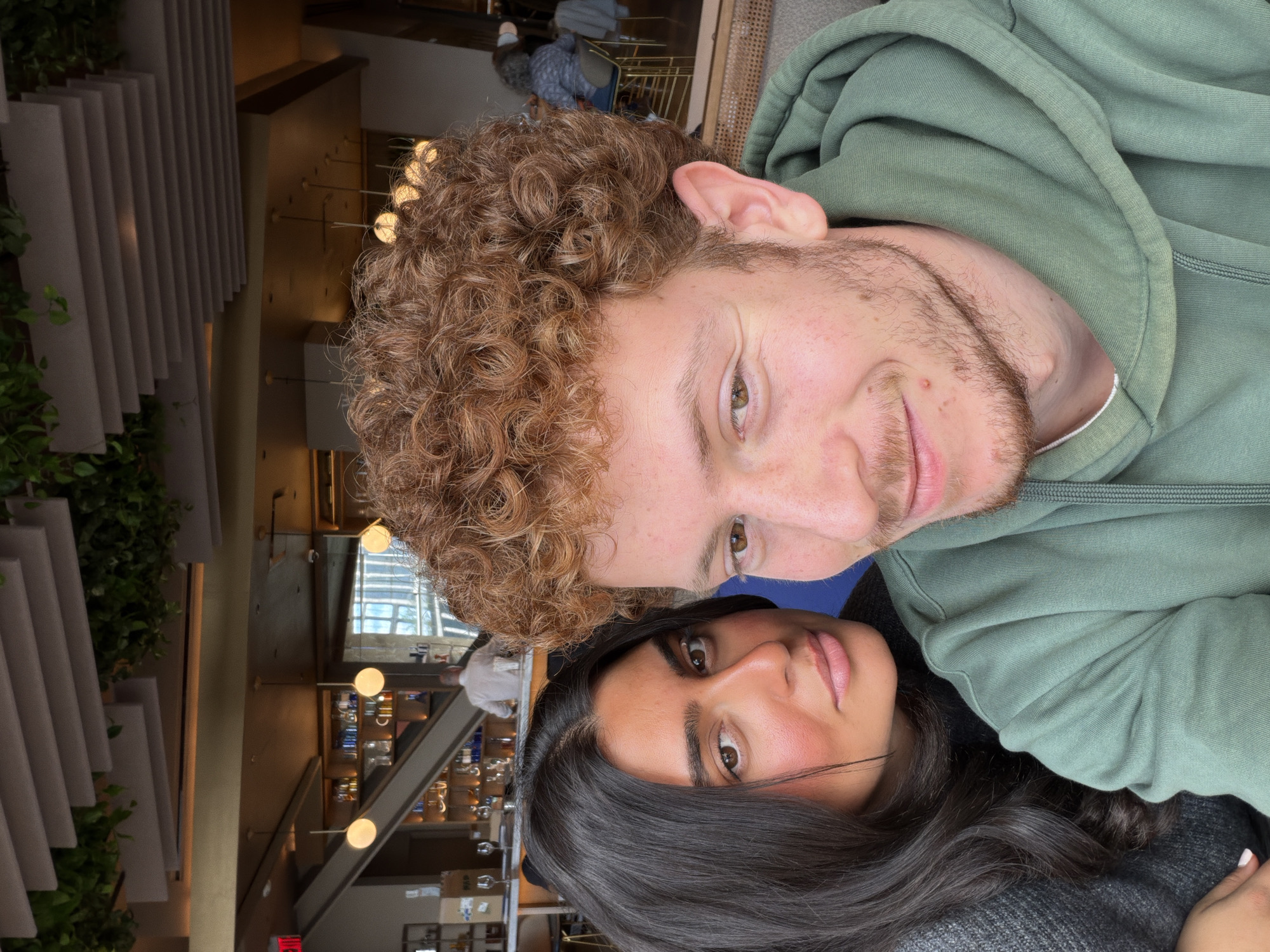 A couple is posing for a close-up selfie. The man has curly red hair, hazel eyes, and a light beard. He is wearing a green hooded sweatshirt. The woman has long dark hair and brown eyes. They appear to be inside a restaurant or bar in New York City. Tables, chairs, and other patrons are visible in the background. A staircase is visible on the left.