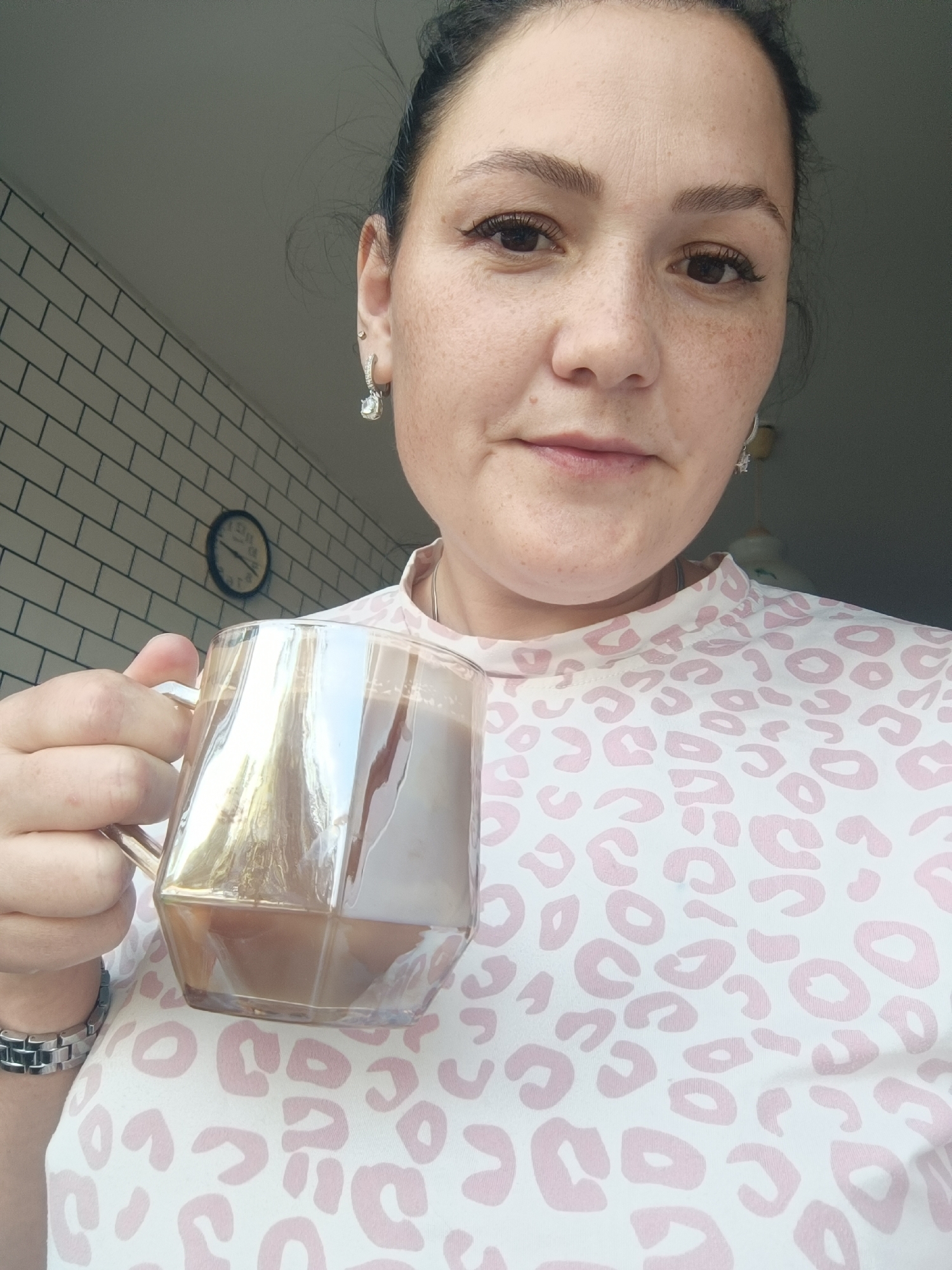 A woman is holding a clear glass mug filled with a brown liquid, possibly coffee, in her left hand. She is looking towards the camera, with her face angled slightly to her left. She has dark hair pulled back, dark eyebrows, and is wearing dangling earrings. She is also wearing a light-colored t-shirt with a pink leopard print pattern. On her left wrist, she has a silver watch with a metal band. In the background, there is a tiled wall and a round clock on the wall. This scene appears to be indoors, possibly in a home or cafe, and there is no indication that this location is in Chaikovsky, Russia.